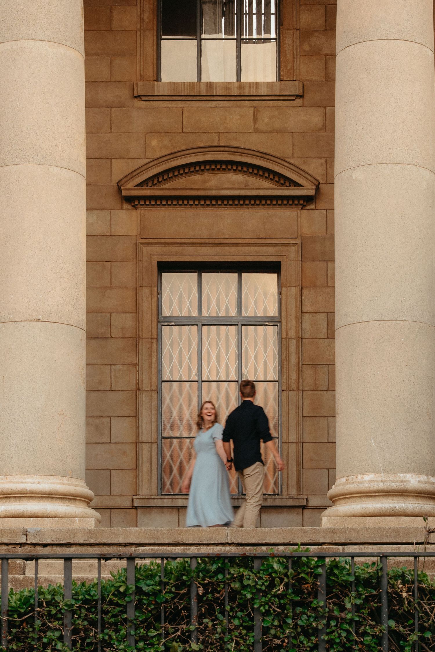 A couple stands between elegant stone columns in front of a historic building with diamond-paned windows.