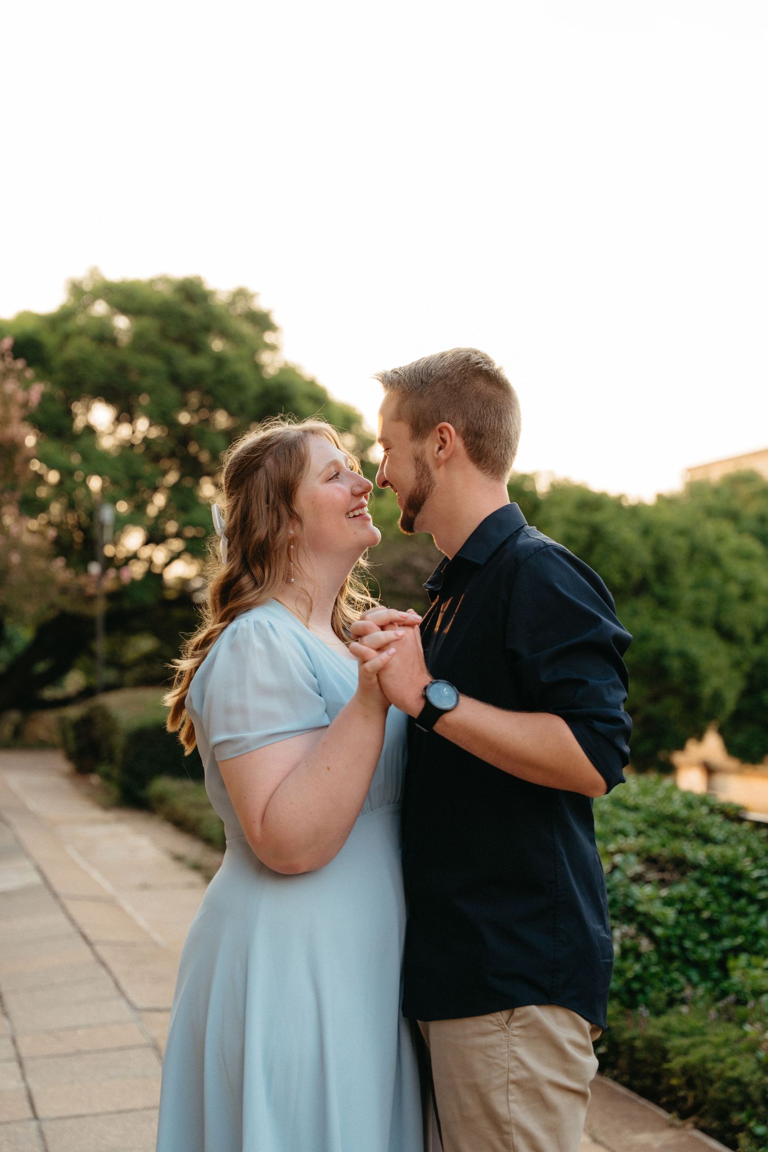 A couple shares a romantic moment while dancing together on a stone path surrounded by greenery at sunset.