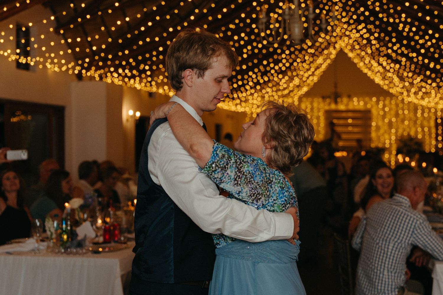 A couple shares a romantic dance under twinkling string lights at an indoor wedding reception.