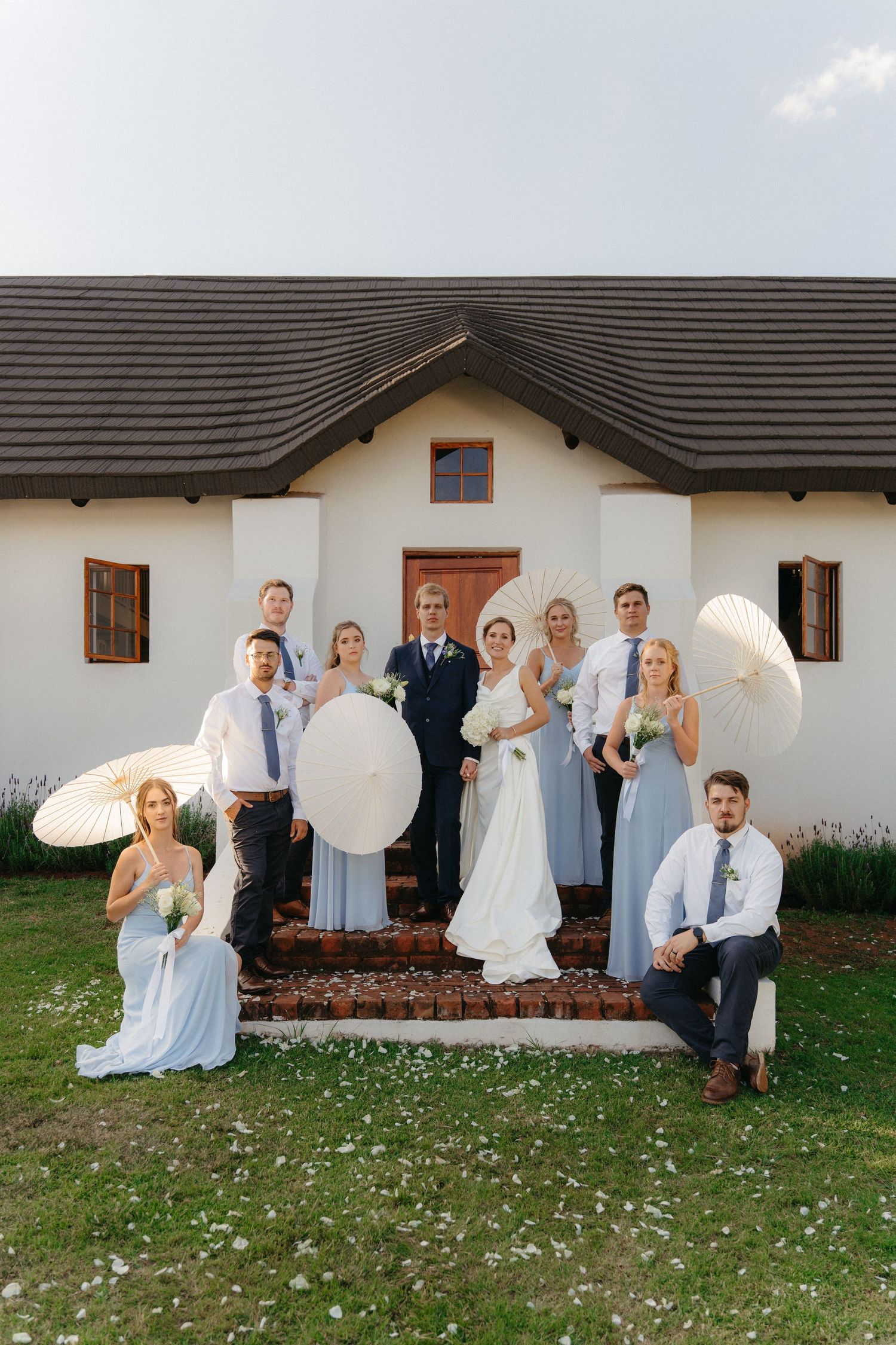 A wedding party poses playfully in a sequence of photos outside a white building with a dark roof.