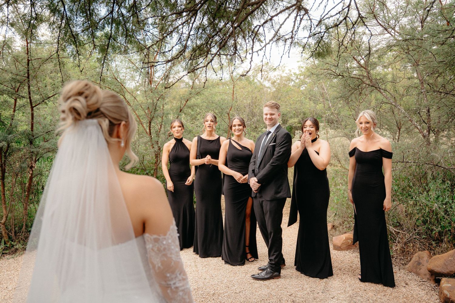 Wedding party in black formal attire stands outdoors in a desert garden setting during a ceremony.