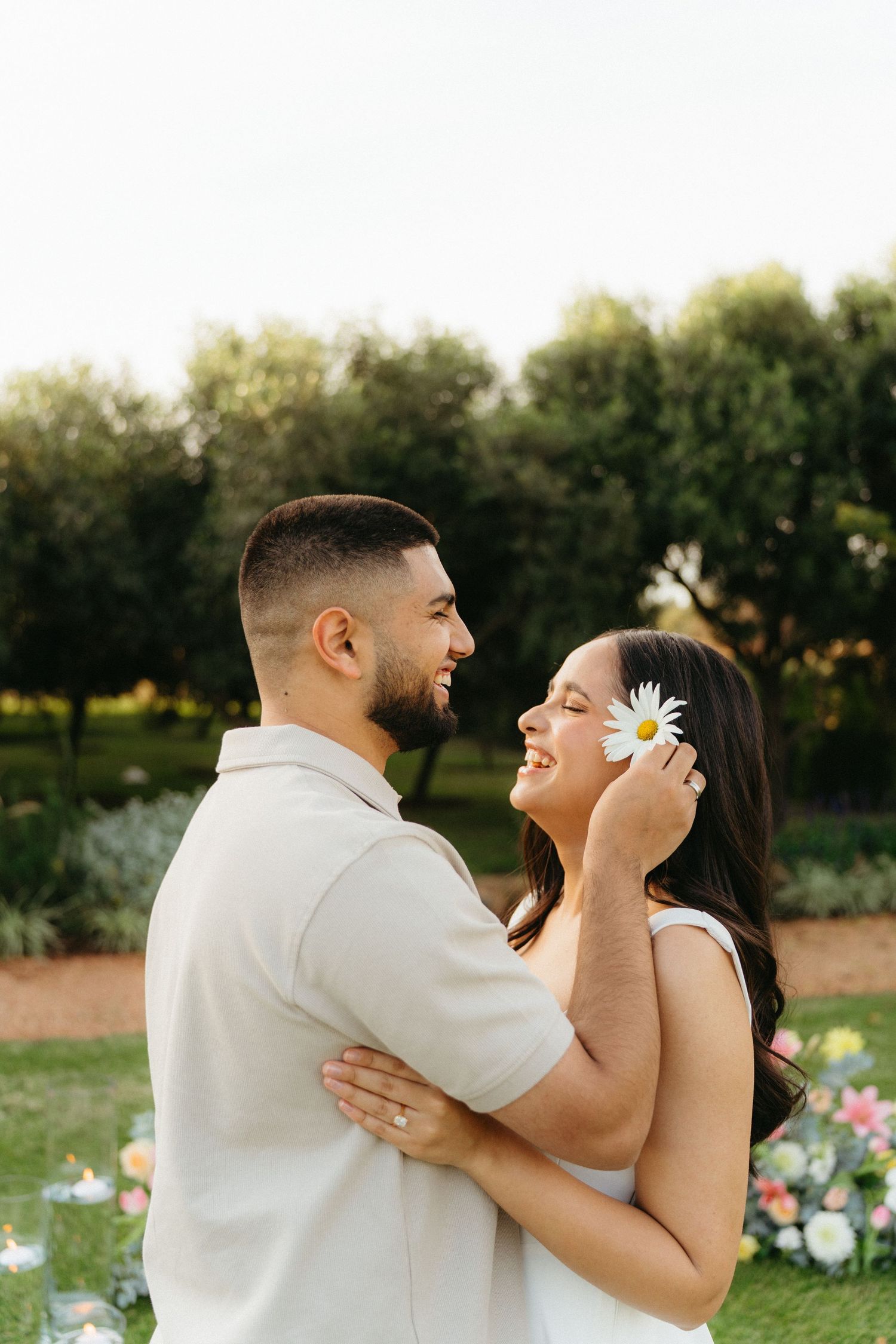 Couple smiles at each other at Ground the Venue, woman holds daisy flower near face, romantic moment.