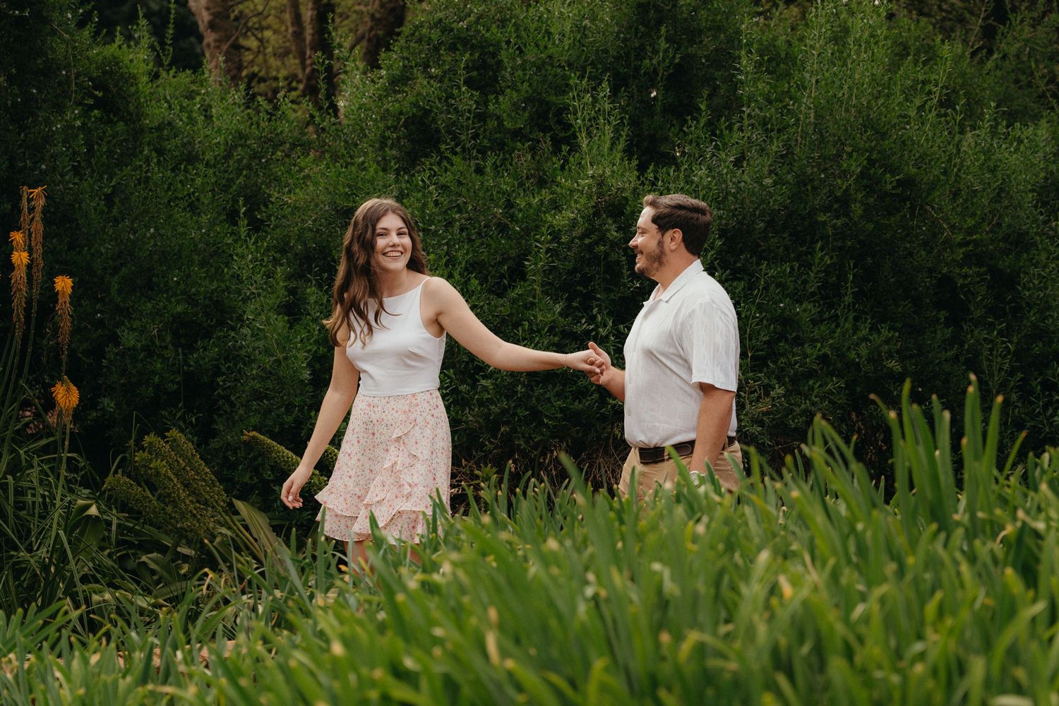Couple holding hands and smiling while walking through lush green garden, woman in white top and floral skirt.