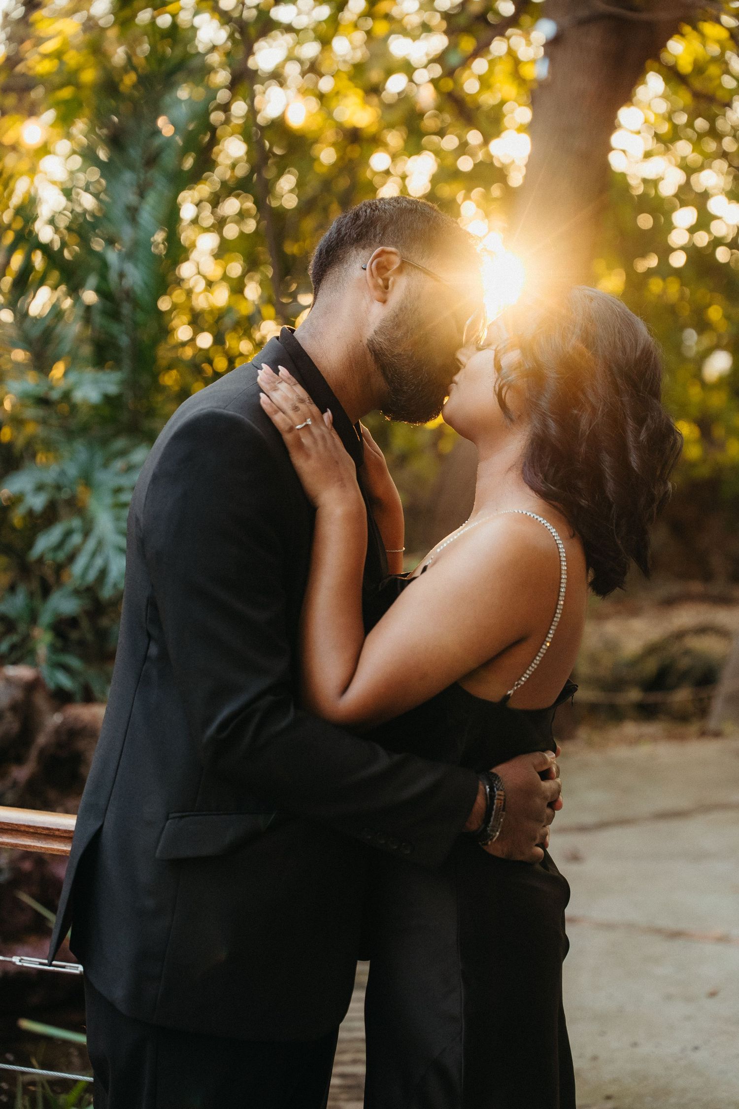 Couple in black outfits sharing a romantic kiss at golden hour with sunlight glowing through lush green trees.
