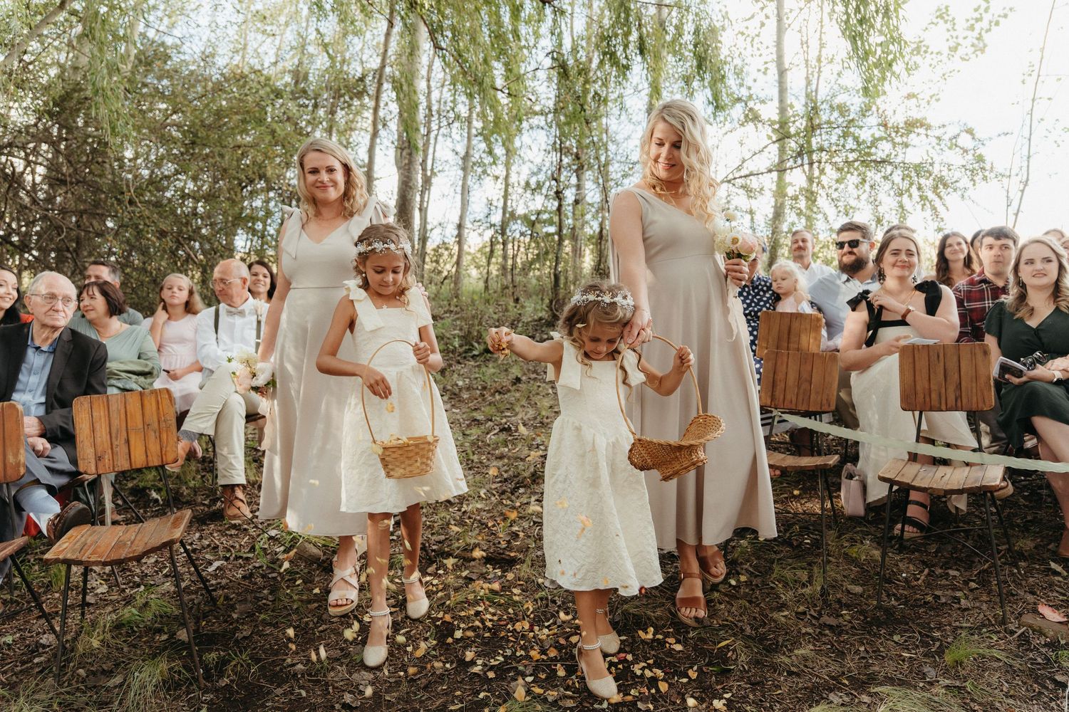 Two flower girls in white dresses walk down an outdoor woodland wedding aisle with bridesmaids in beige gowns.