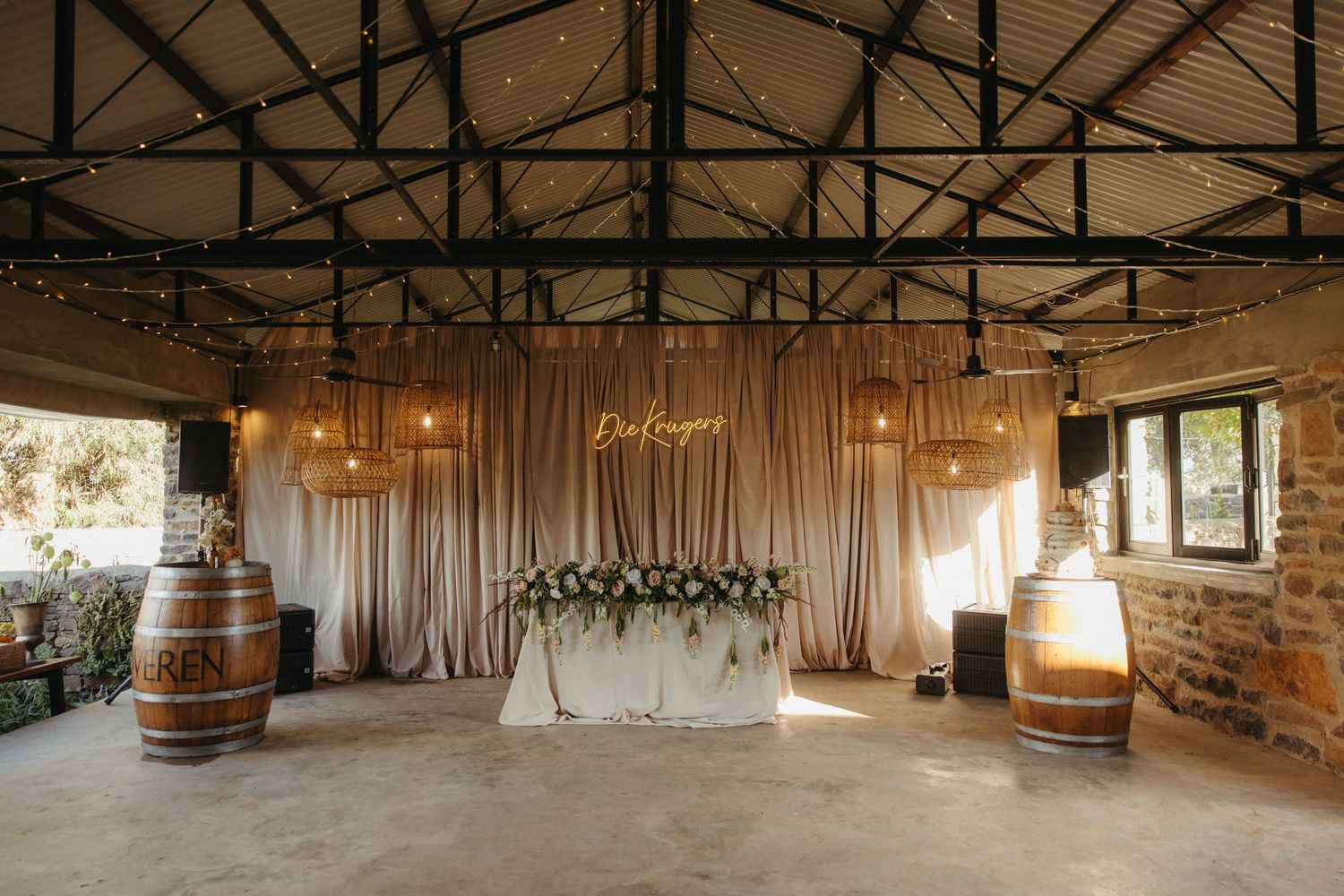 Elegant wedding reception venue with draped fabric backdrop, floral centerpiece, wine barrel decor, and warm string lights overhead.