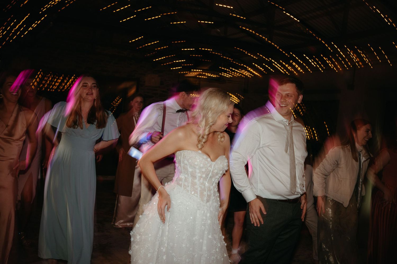 Bride in white gown dancing joyfully with guests at a wedding reception under warm string lights.