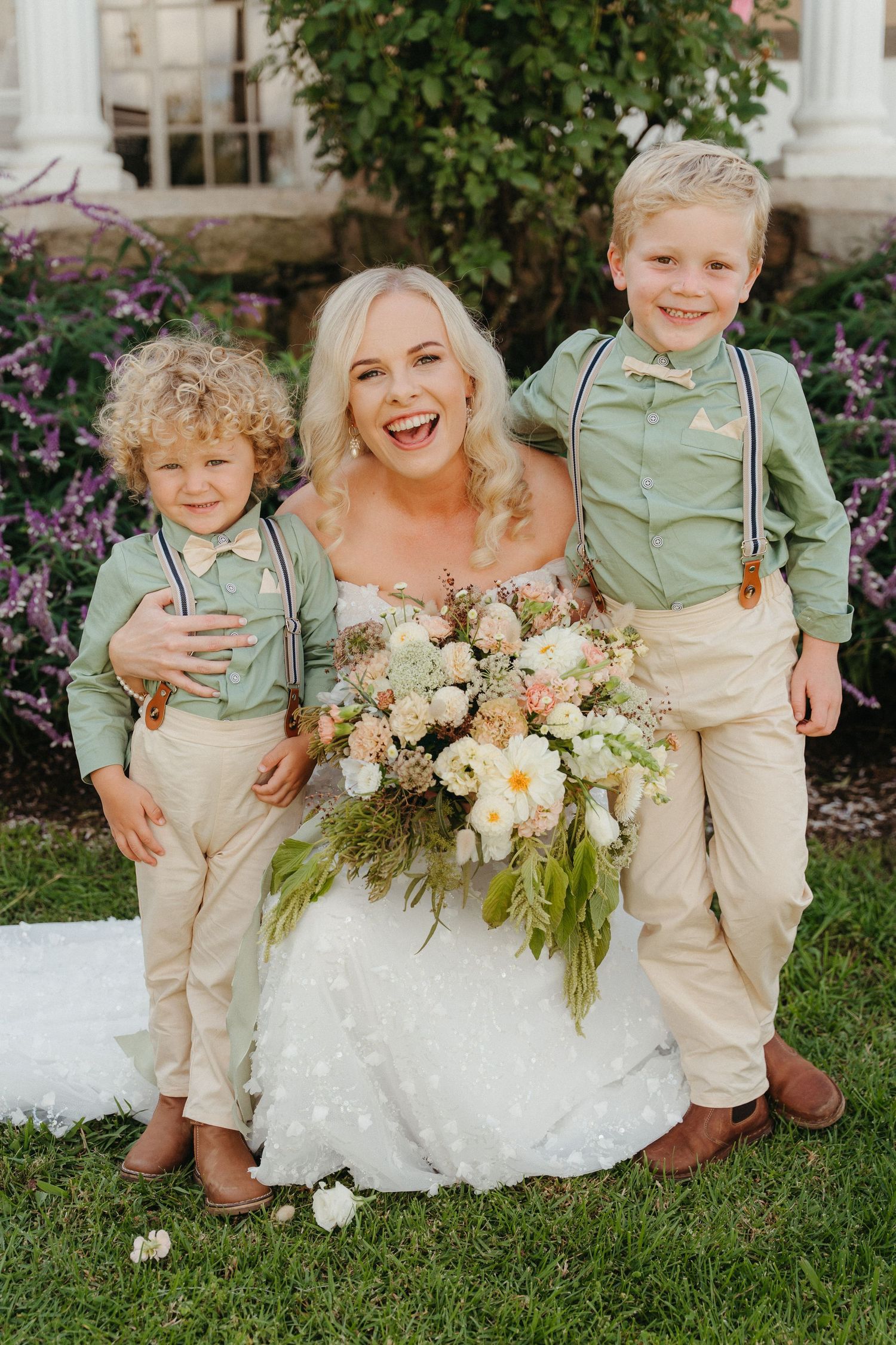 Smiling bride in white gown holds floral bouquet while posing with two young boys in green shirts and suspenders outdoors.
