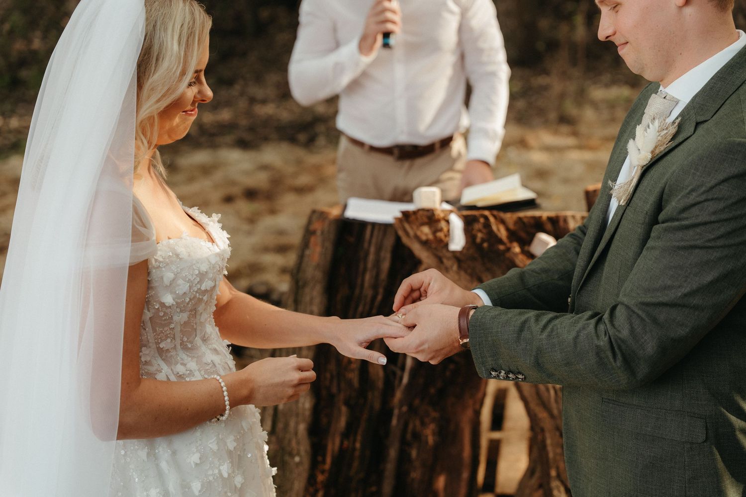 Bride and groom exchanging rings during an outdoor rustic wedding ceremony with a officiant in background.
