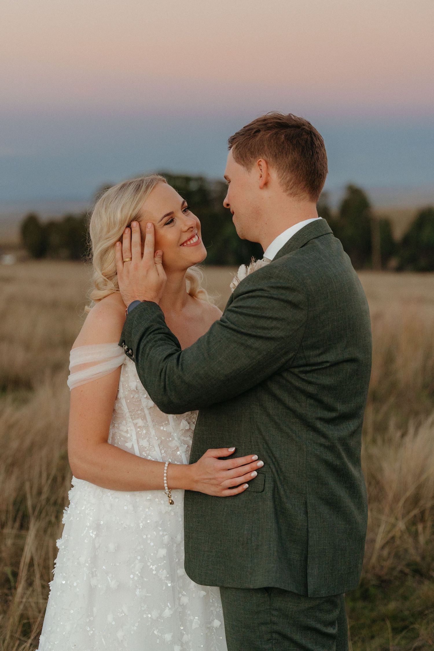 A groom in a green suit tenderly cups his bride's face during a romantic golden hour outdoor wedding portrait.