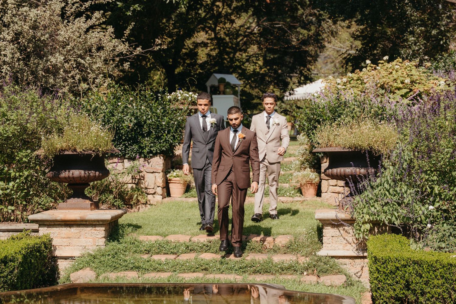 Three groomsmen in brown and beige suits walk through a lush garden pathway at an outdoor wedding ceremony.
