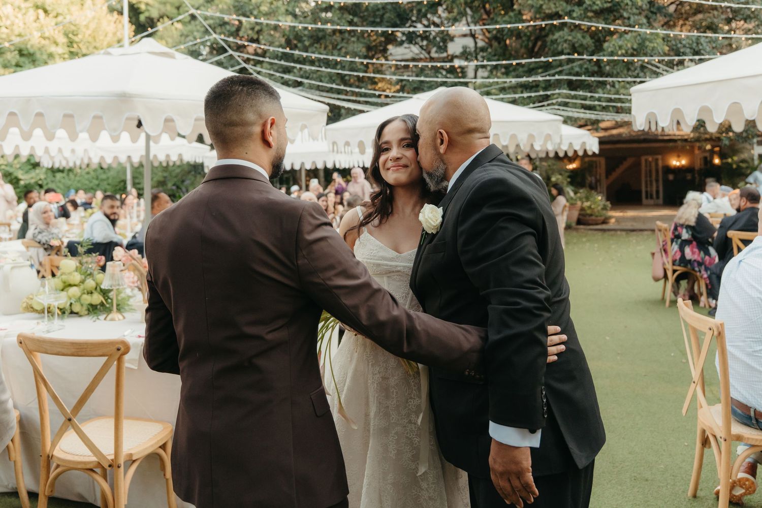 Bride in white gown greeted by guests at outdoor wedding reception with string lights and white tents.