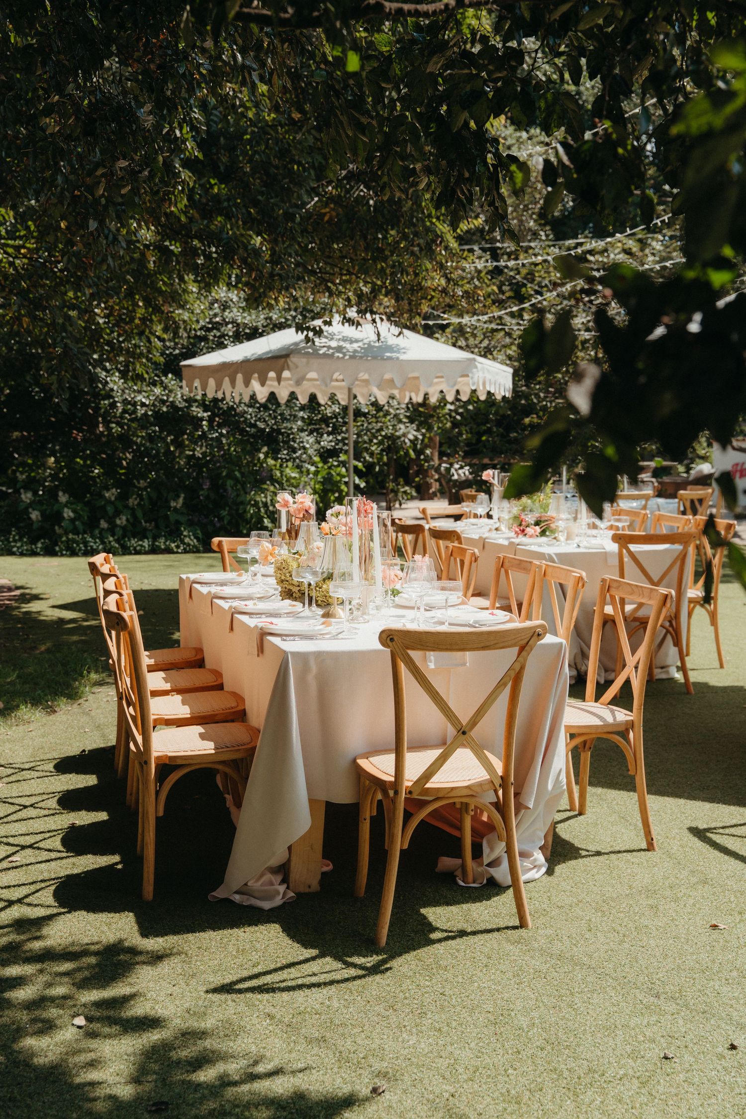 Elegant outdoor wedding reception with wooden cross-back chairs, white linen tables, and a scalloped umbrella on a green lawn.