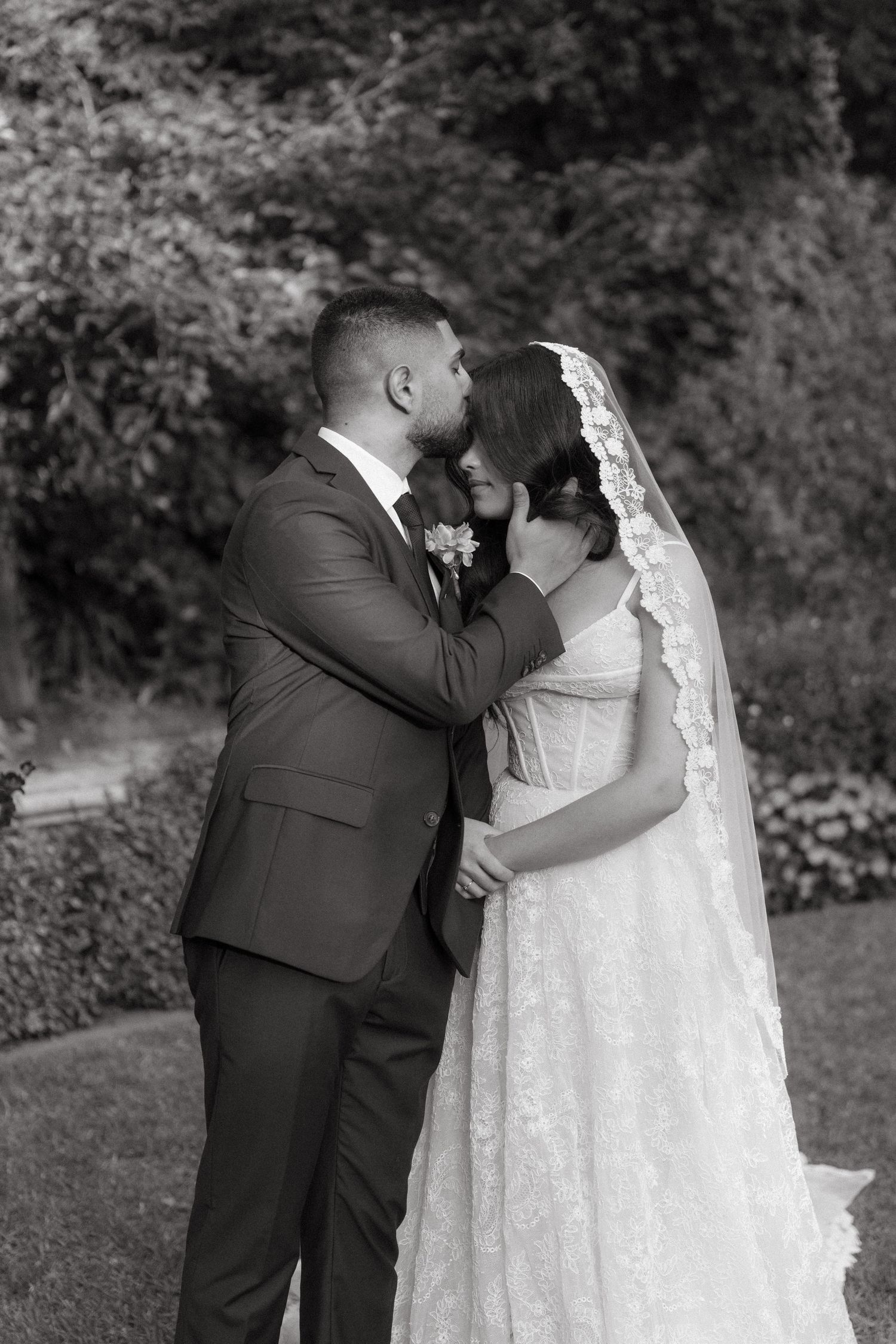 Black and white photo of a groom kissing his bride on the forehead in a lush garden setting.