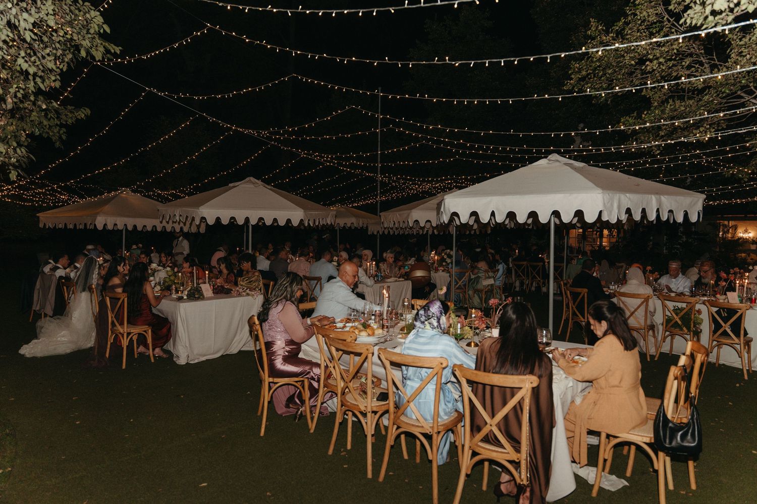 Outdoor wedding reception at night with string lights, white tent, and guests dining at wooden tables on green grass.