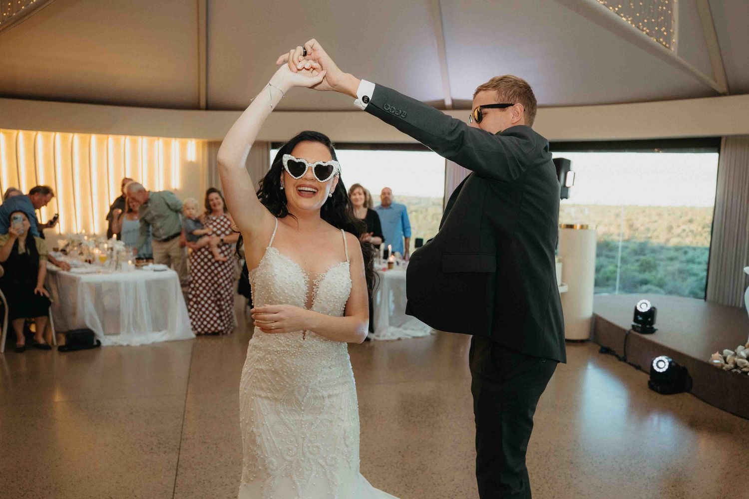 Bride in white gown and heart sunglasses dances with groom in black suit at elegant wedding reception.