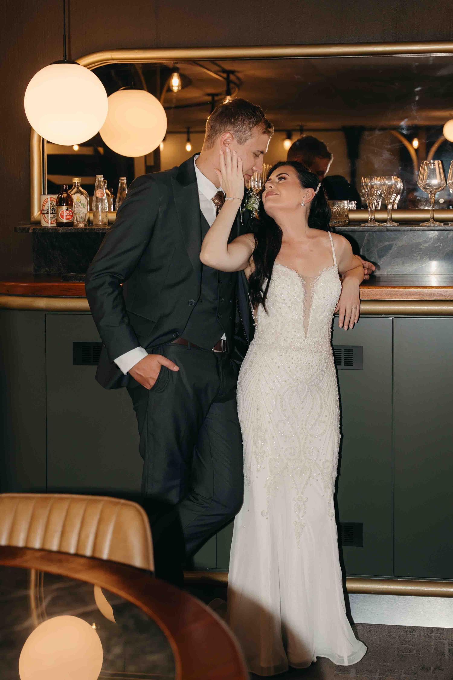 A bride in a beaded gown and groom in a dark suit share a romantic moment at an elegant bar venue.