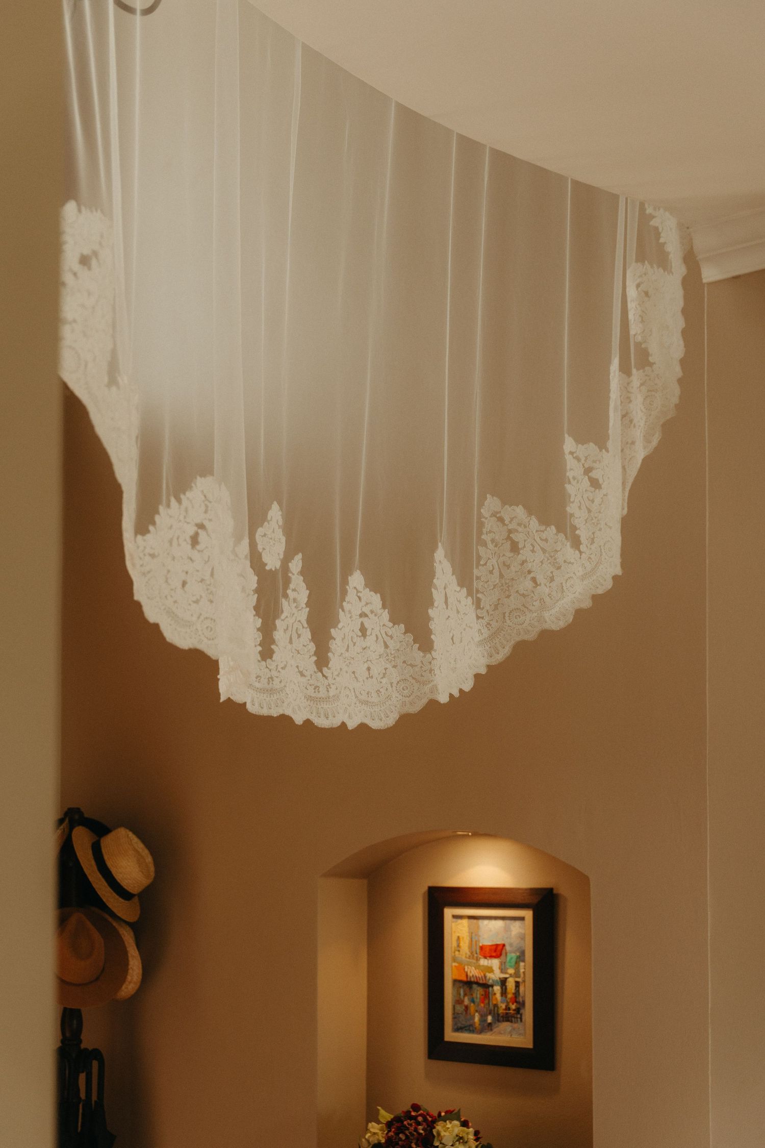 White lace wedding veil displayed on ceiling above a warm-toned hallway with framed artwork and decorative hats.