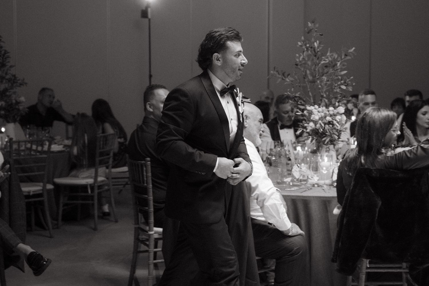 Man in tuxedo stands attentively at elegant wedding reception dinner in black and white photo.
