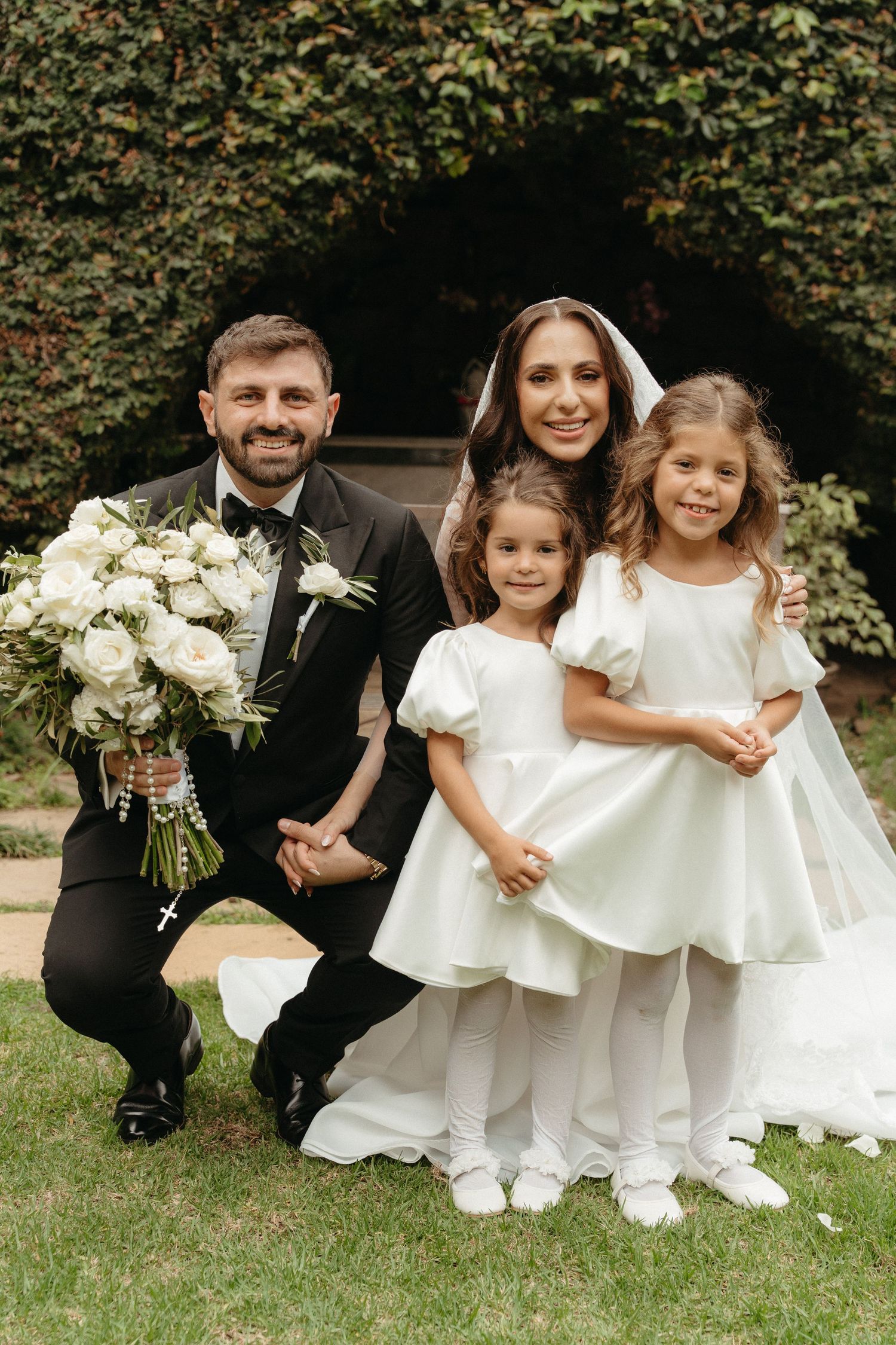 A smiling groom in a black tuxedo kneels beside two flower girls in white dresses, with the bride standing behind them outdoors.