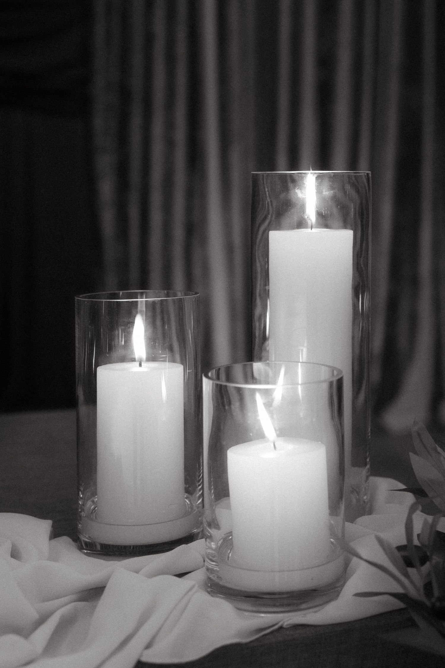 Three lit pillar candles in glass hurricane vases arranged on white fabric in elegant black and white photography.