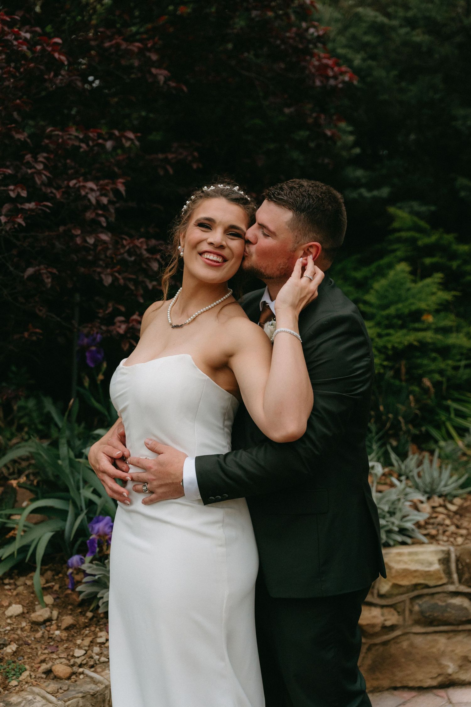 Smiling bride in a strapless white gown and pearl necklace is embraced and kissed on the cheek by her groom in a garden wedding portrait.
