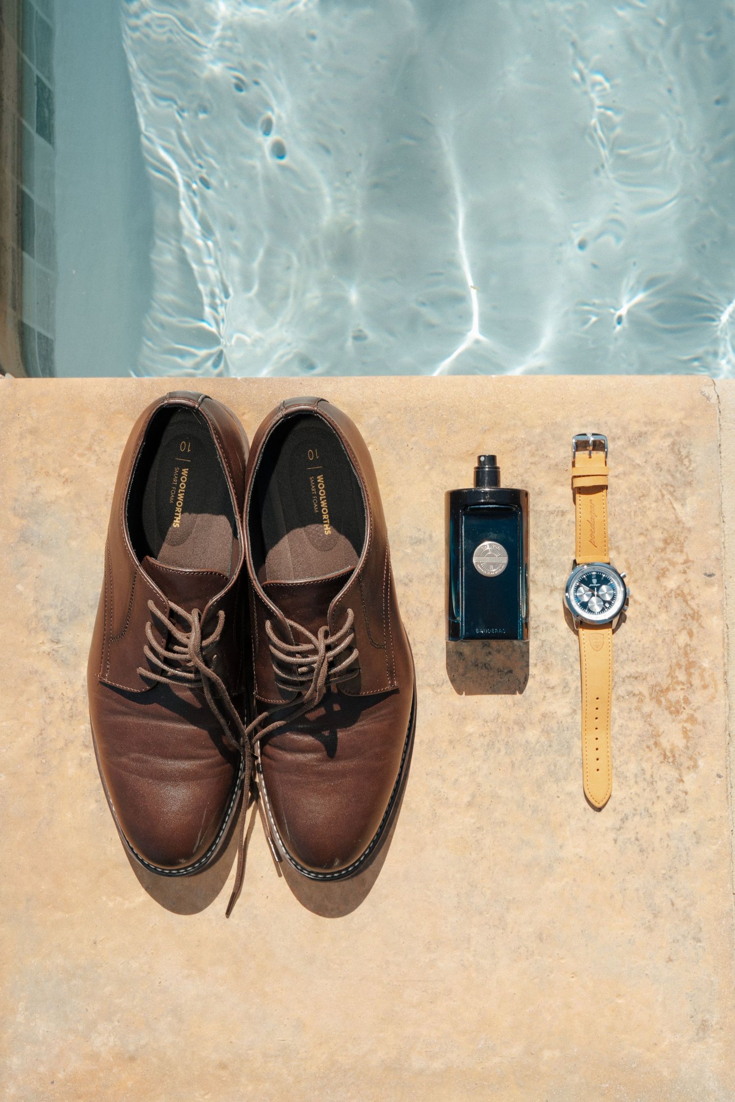 Brown leather derby shoes, a dark cologne bottle and a tan leather strap watch arranged on a stone pool ledge beside sparkling water.