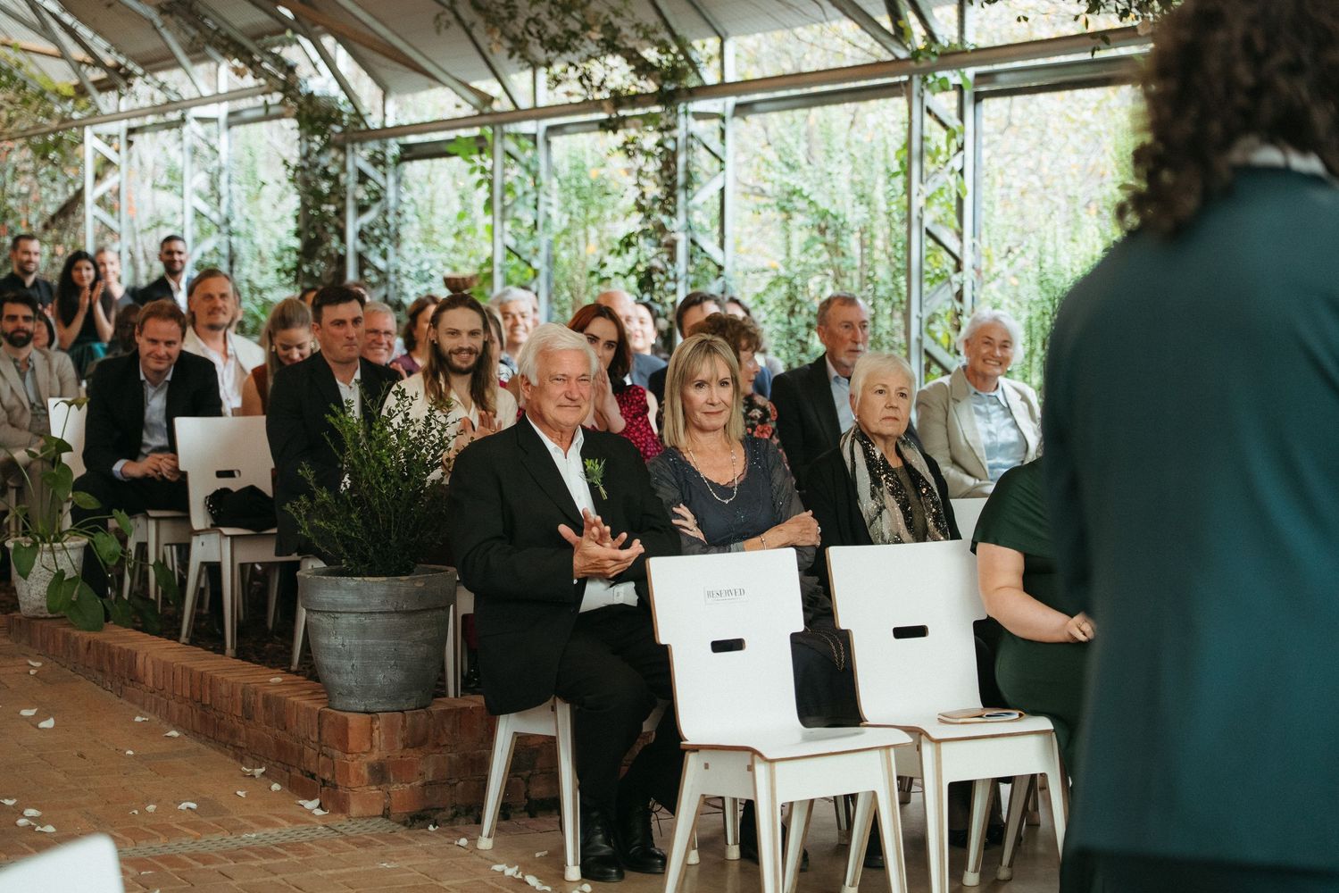 Guests seated in white chairs at an elegant greenhouse wedding ceremony, surrounded by lush greenery and natural light.