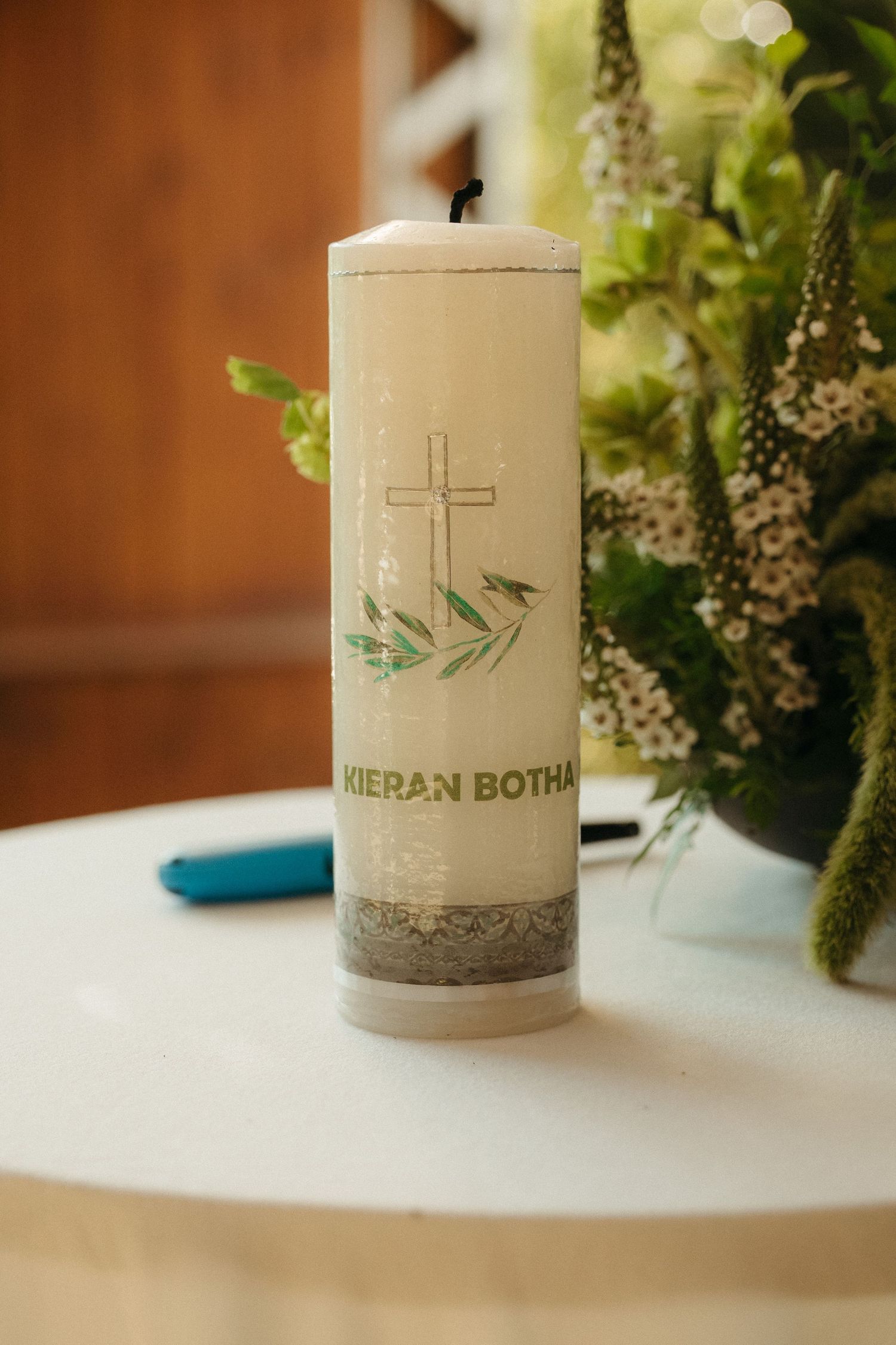 Personalized white baptism candle for Kieran Botha featuring a cross and olive branch design, displayed on a white table.
