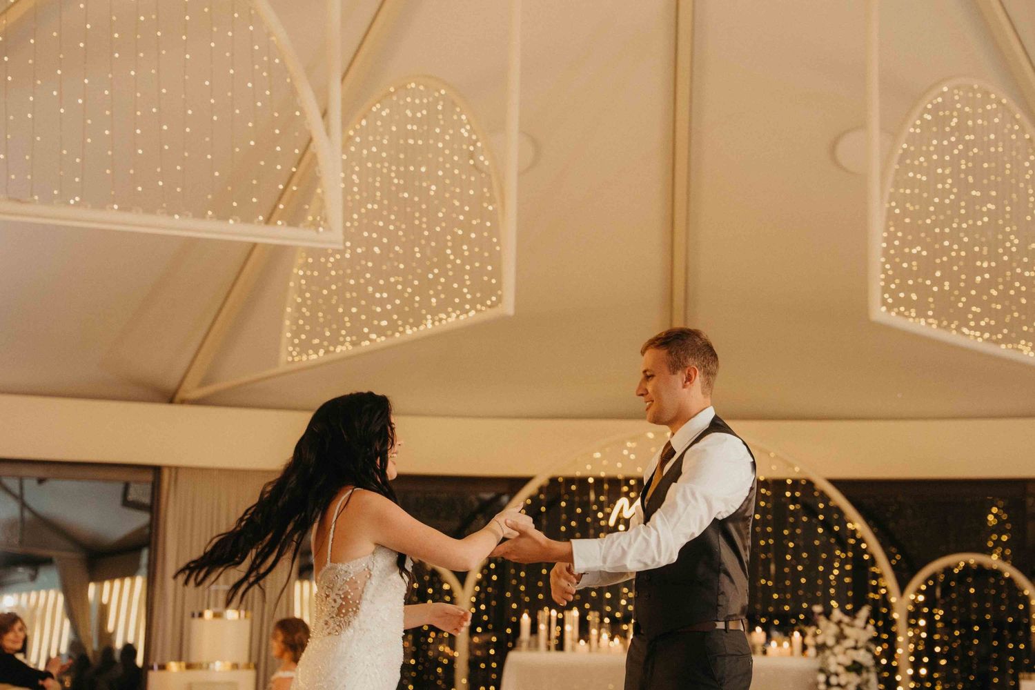 A bride and groom share their first dance under warm fairy lights in an elegant wedding reception venue.