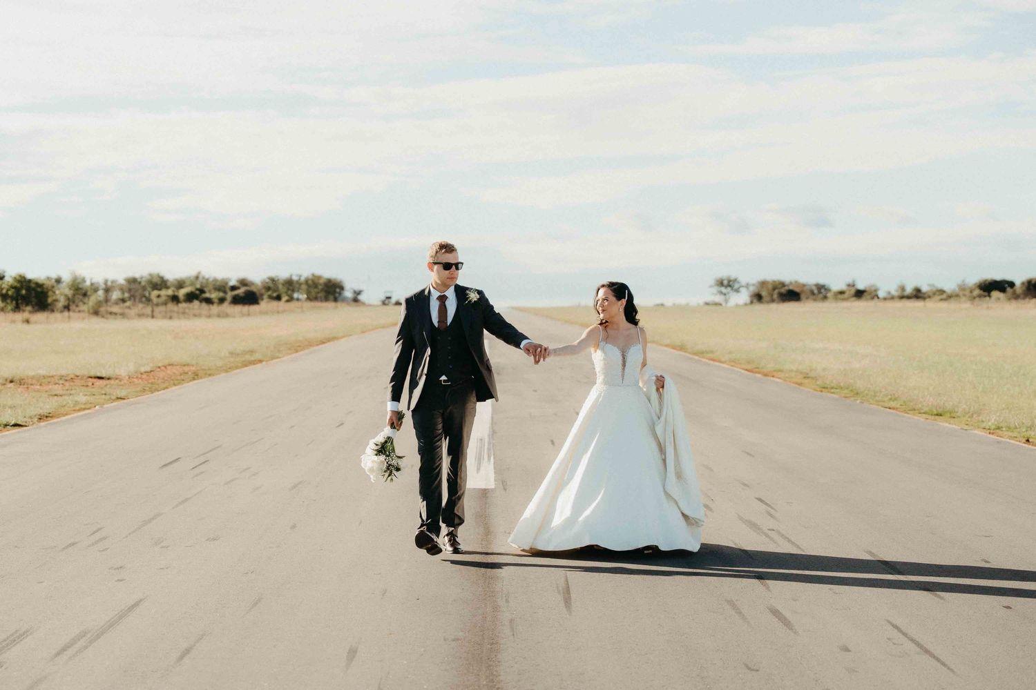 Bride and groom walk hand-in-hand down an open runway road on their wedding day under a cloudy sky.
