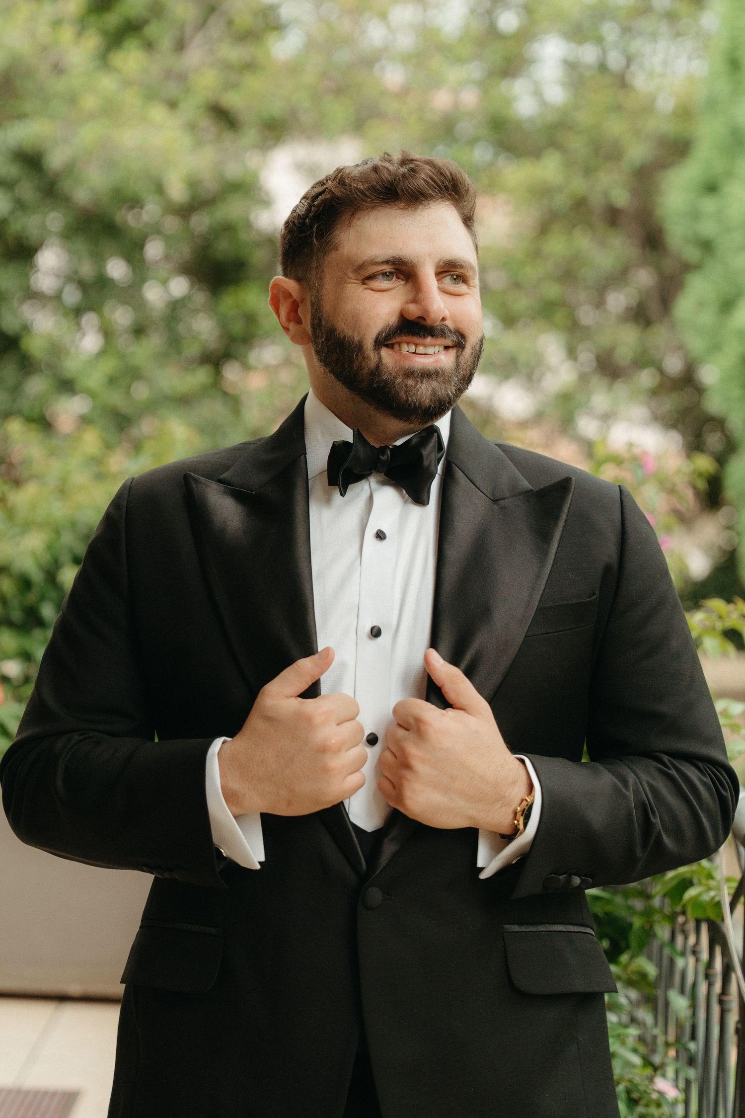 Smiling bearded groom in black tuxedo and bow tie adjusting jacket outdoors with green trees in Johannesburg
