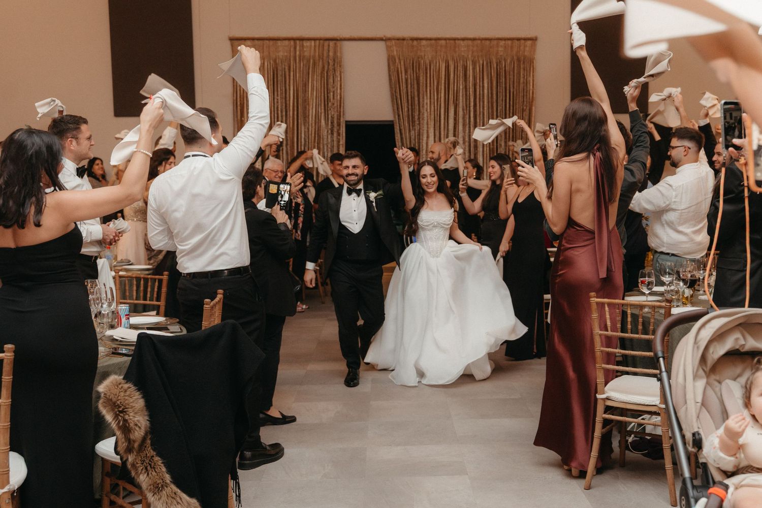 Bride and groom walk through cheering guests waving napkins at elegant wedding reception celebration.