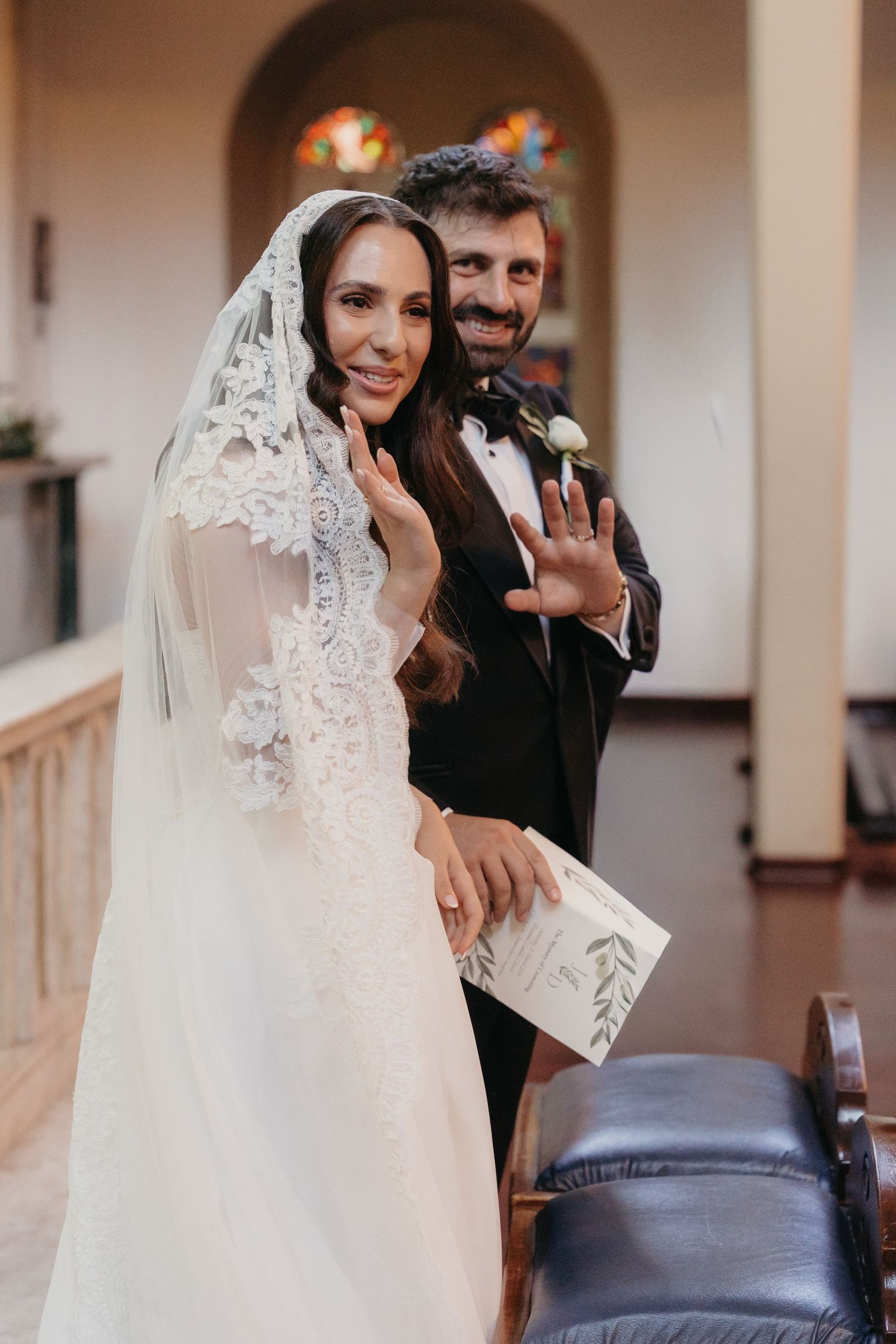 Bride in lace veil and groom in tuxedo showing wedding rings, smiling at camera in elegant church venue.