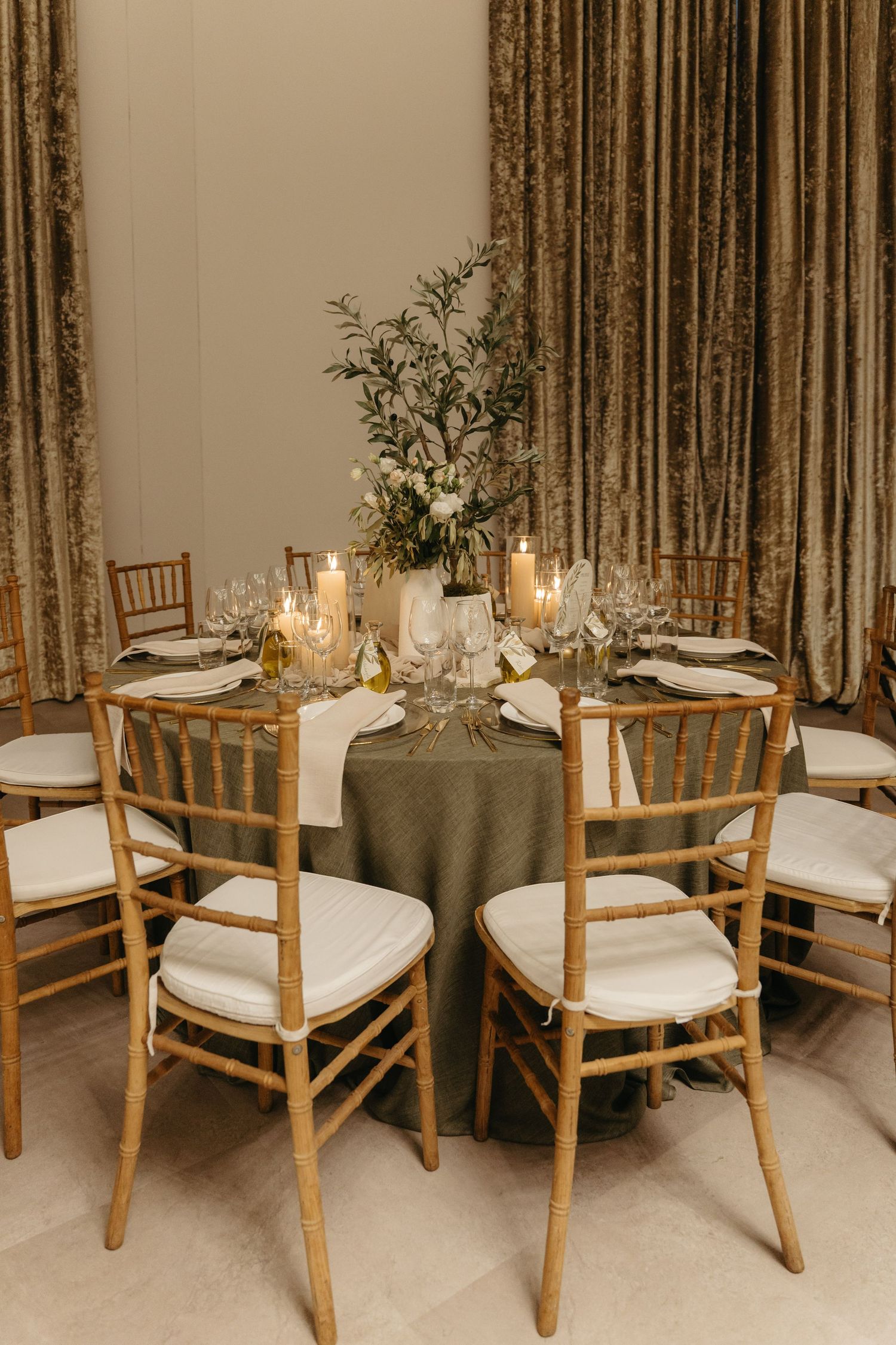 Elegant round wedding reception table with olive green linen, gold chiavari chairs, candles, and floral centerpiece.