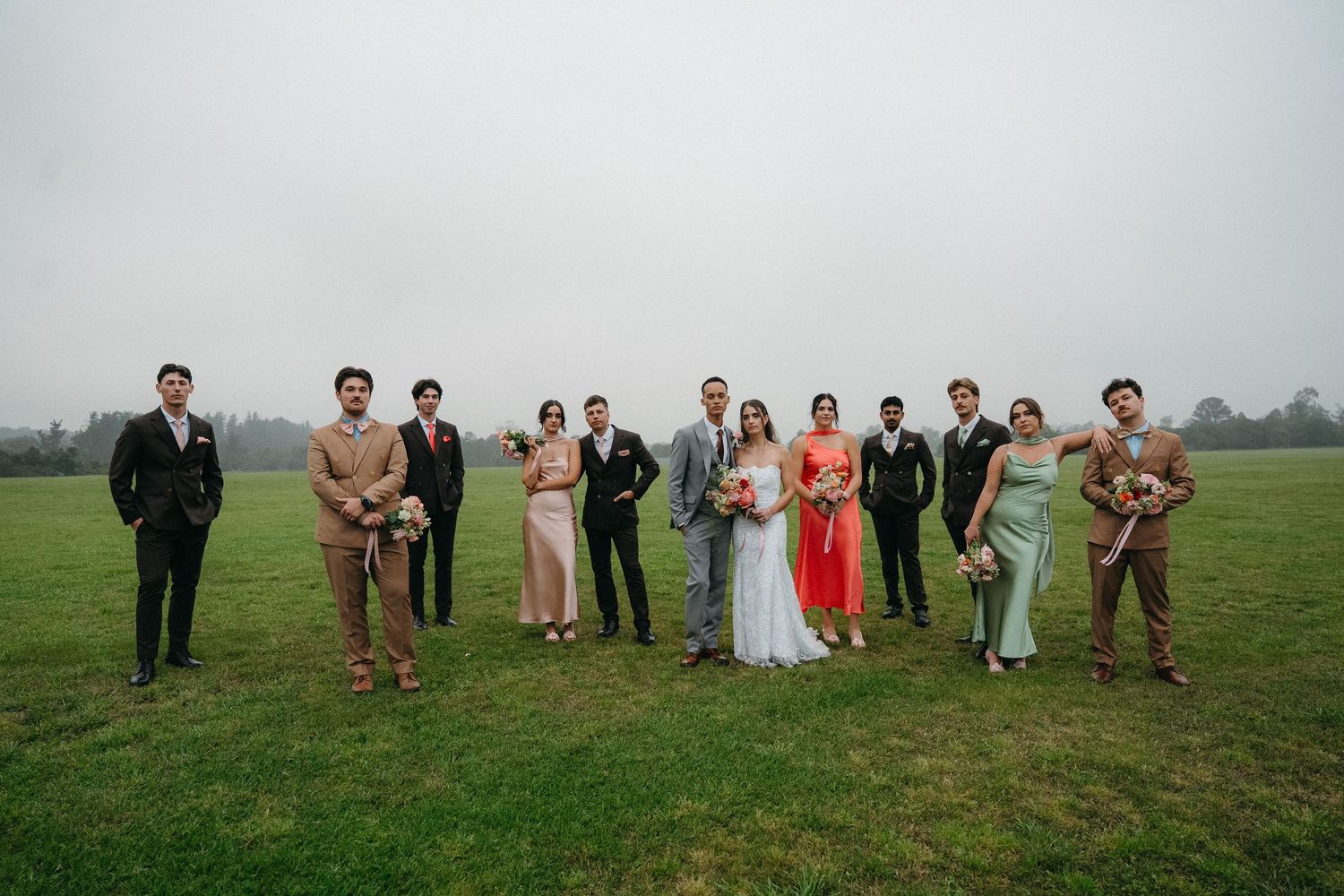 Wedding party poses on misty green field, bride in red, groom in suit, bridesmaids and groomsmen in colorful attire.
