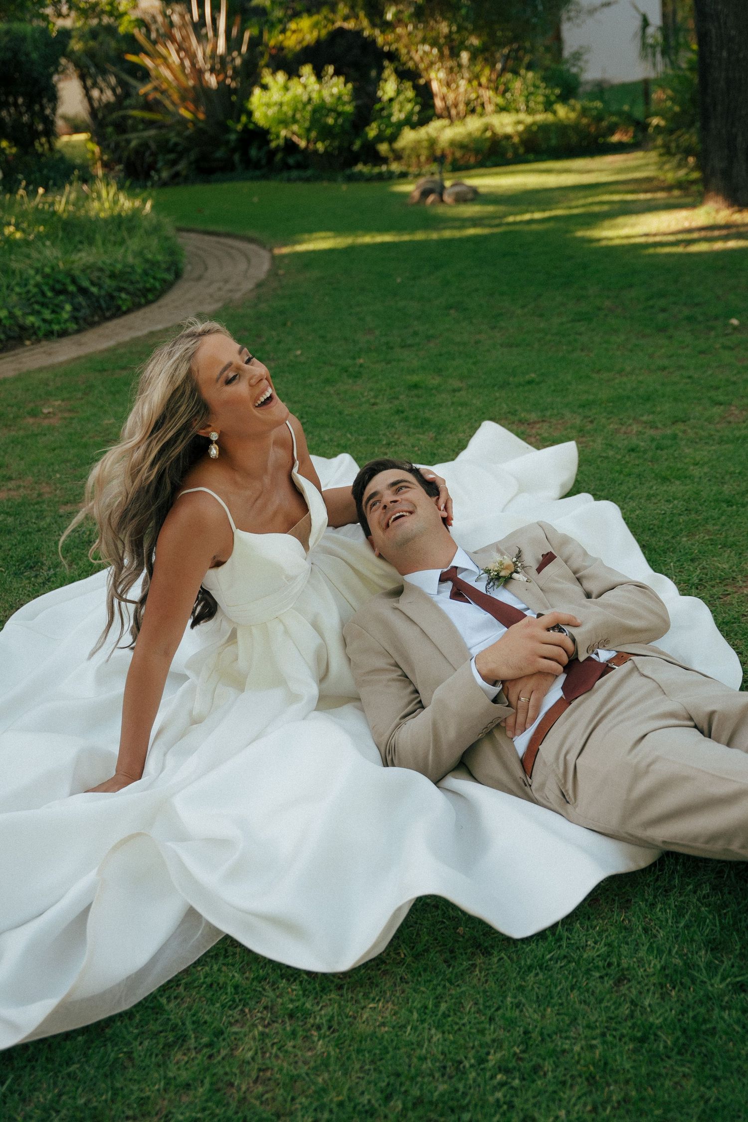 Bride in a white gown and groom in a tan suit laugh together while relaxing on a sunlit green lawn at Duvon Wine Estate.
