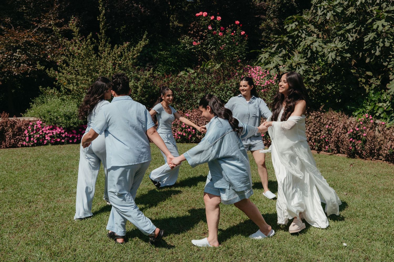 Bride in white gown dancing in a circle with bridesmaids in light blue pajamas on green lawn surrounded by blooming gardens.