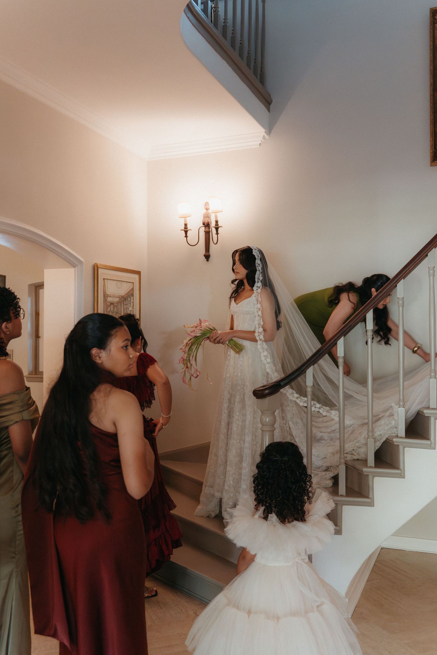 Bride in white lace gown and veil descends staircase holding bouquet, surrounded by bridesmaids in burgundy dresses.