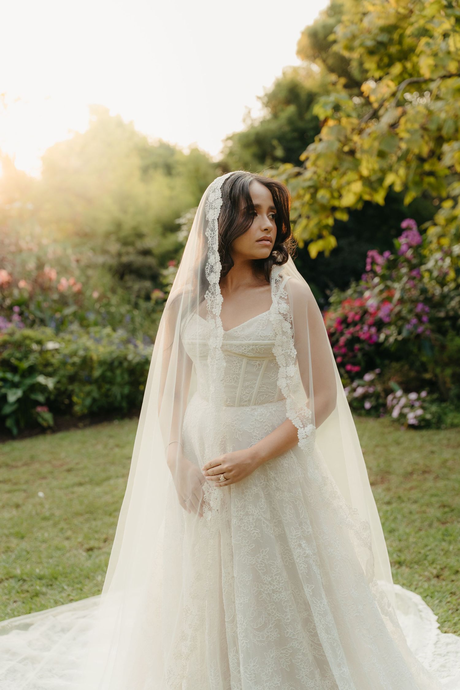 Bride in lace wedding gown and veil standing in sunlit garden with colorful flowers.