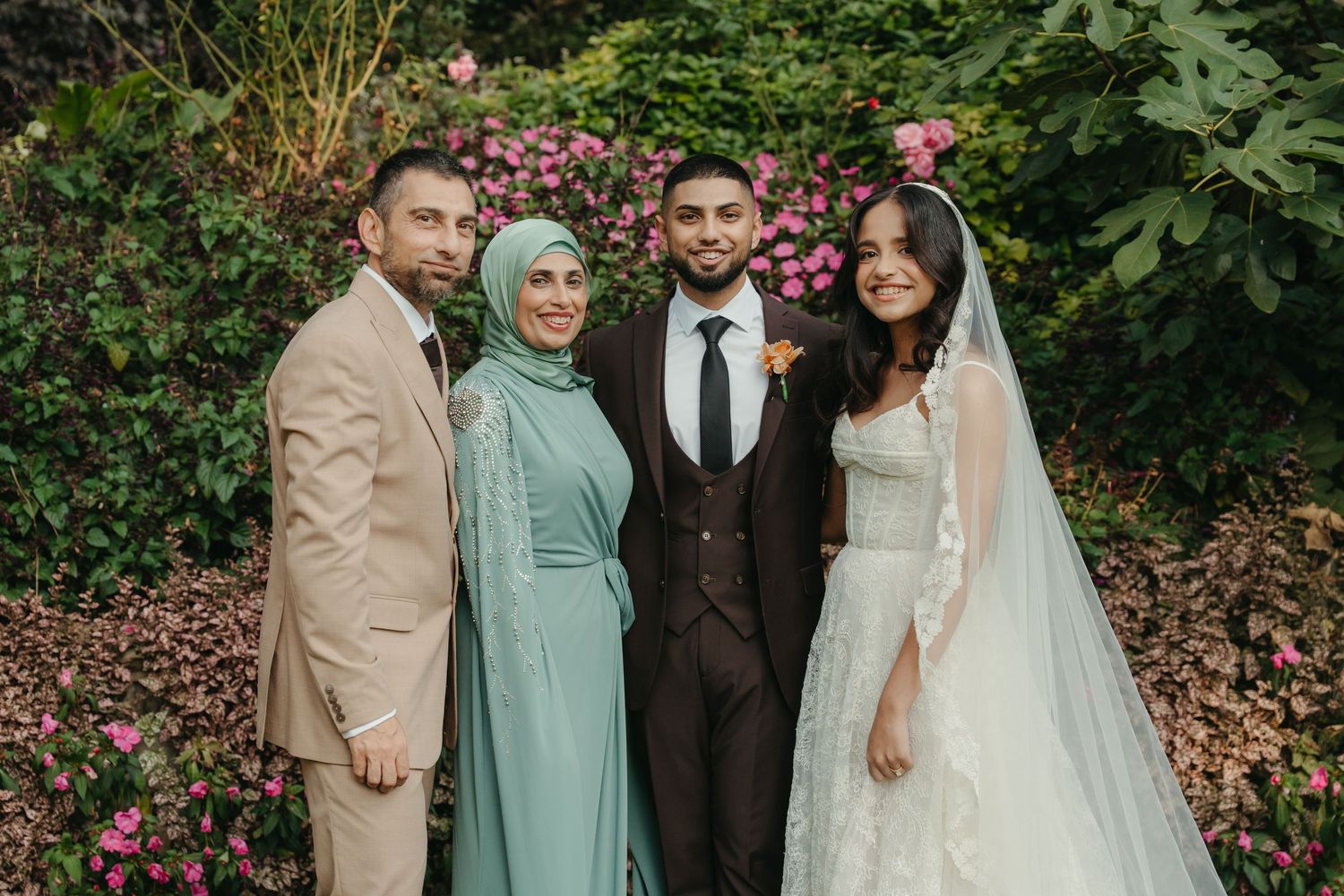 Wedding party of four posing outdoors, bride in white gown, groom in dark suit, guests in sage green hijab and beige suit.