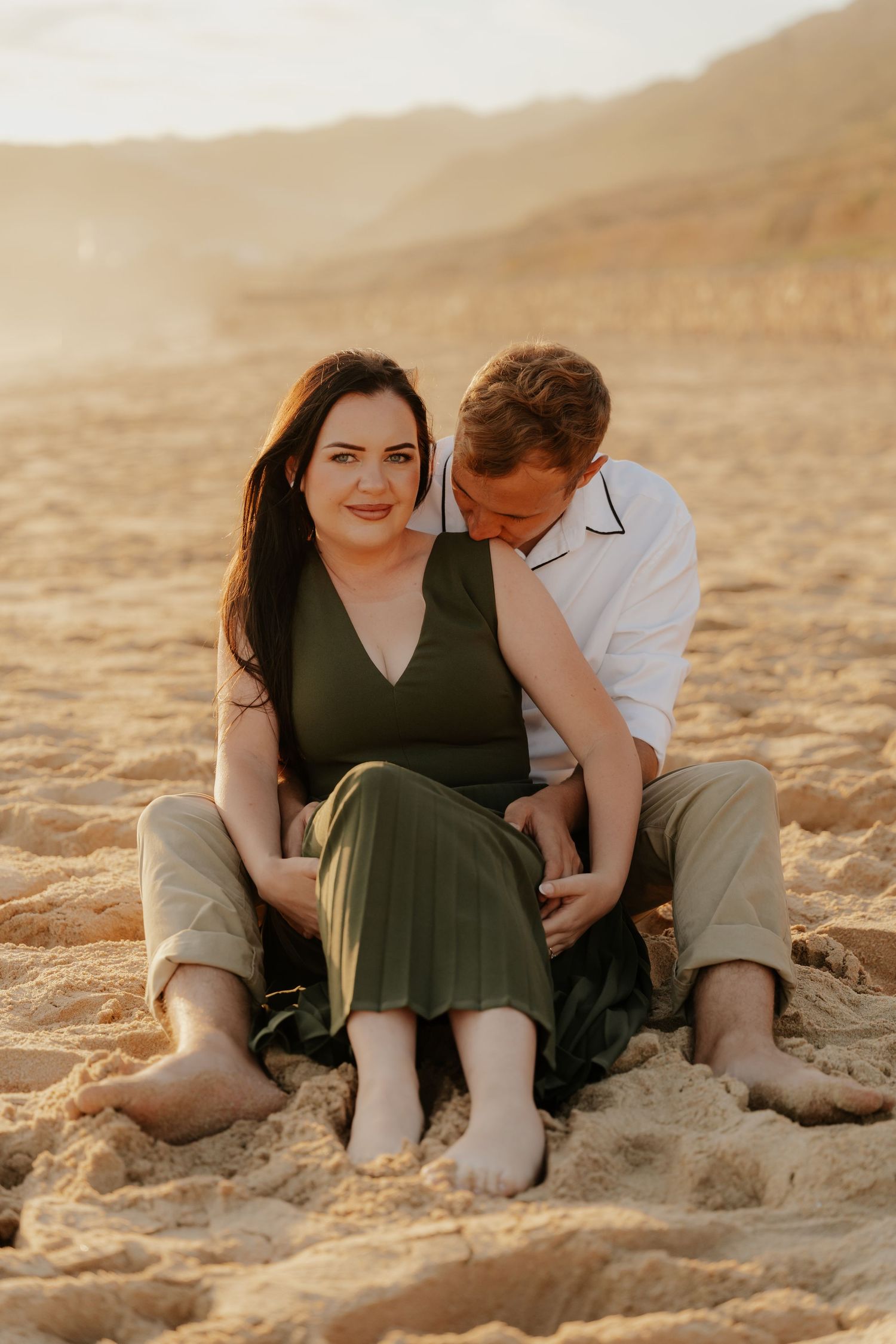 A romantic couple embraces on a sunlit beach in Mossel bay with warm tones and Western Cape mountains visible in the distance.