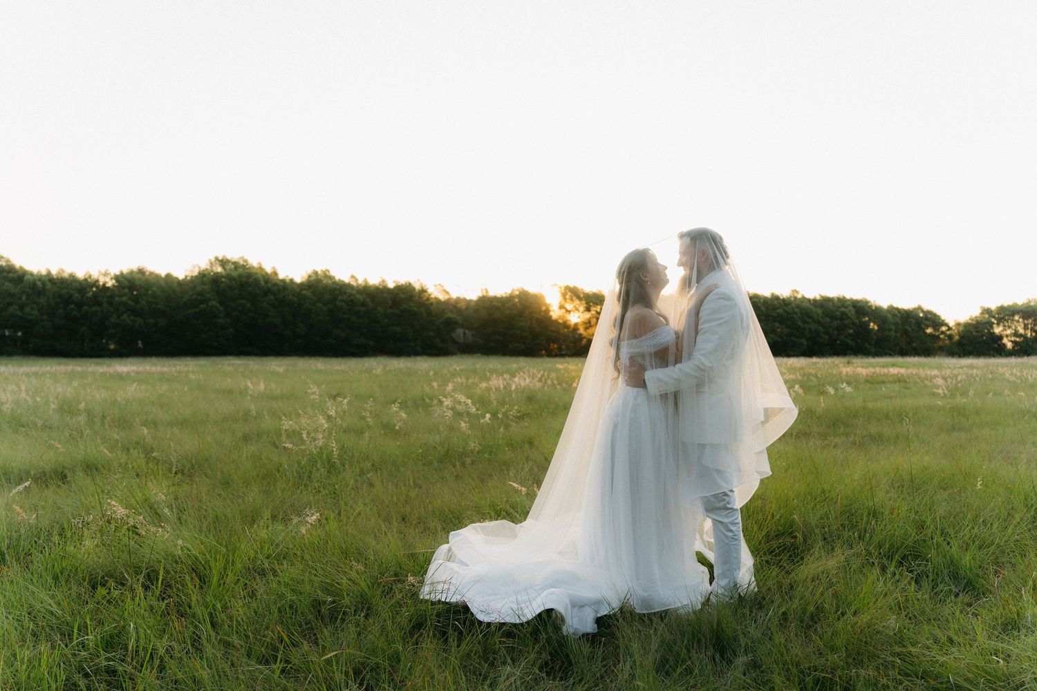 Newlyweds embrace in a serene meadow during sunset with the bride's veil flowing in the breeze.