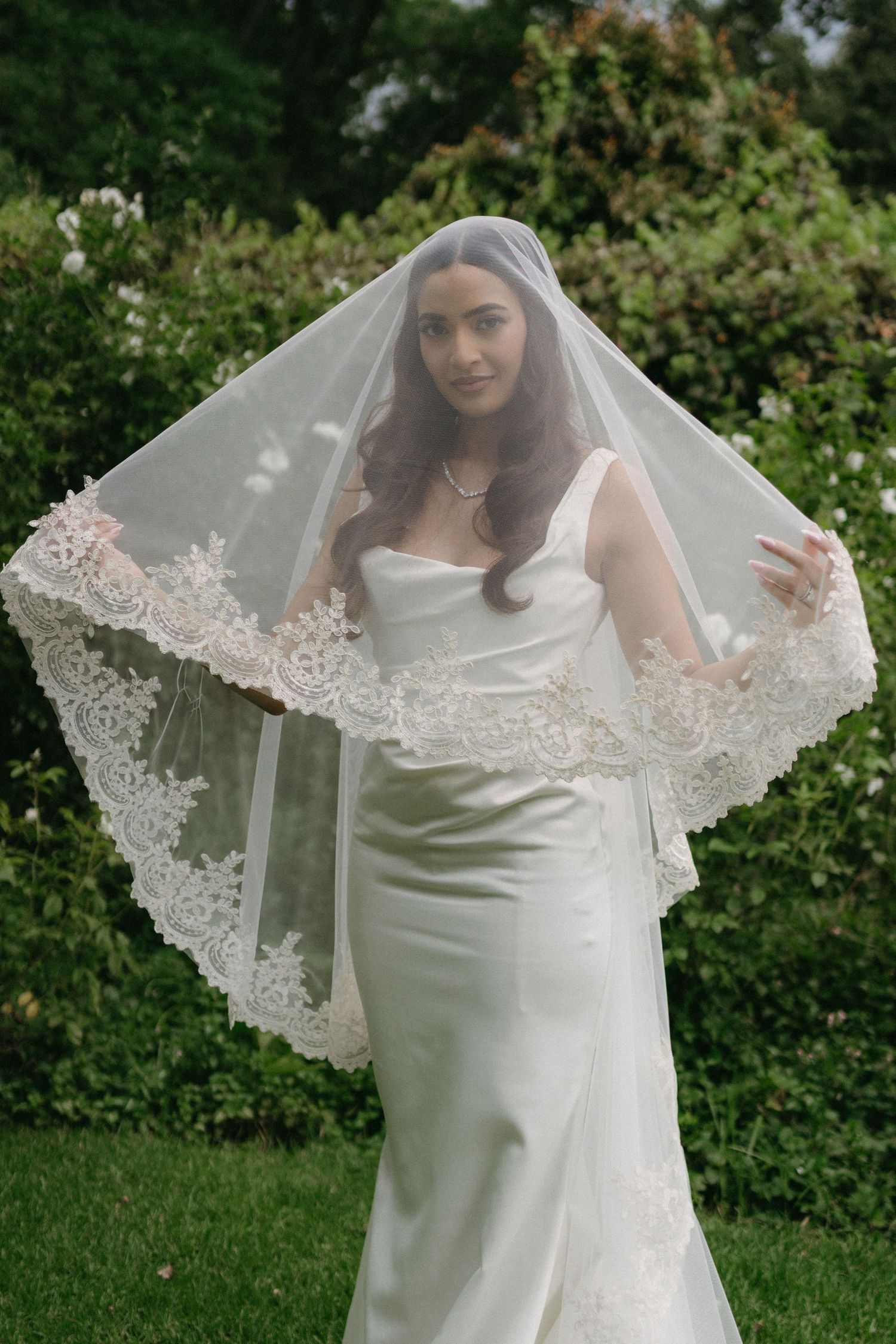 Elegant bride in a form-fitting white wedding dress with a delicate lace-trimmed veil stands in a garden setting.