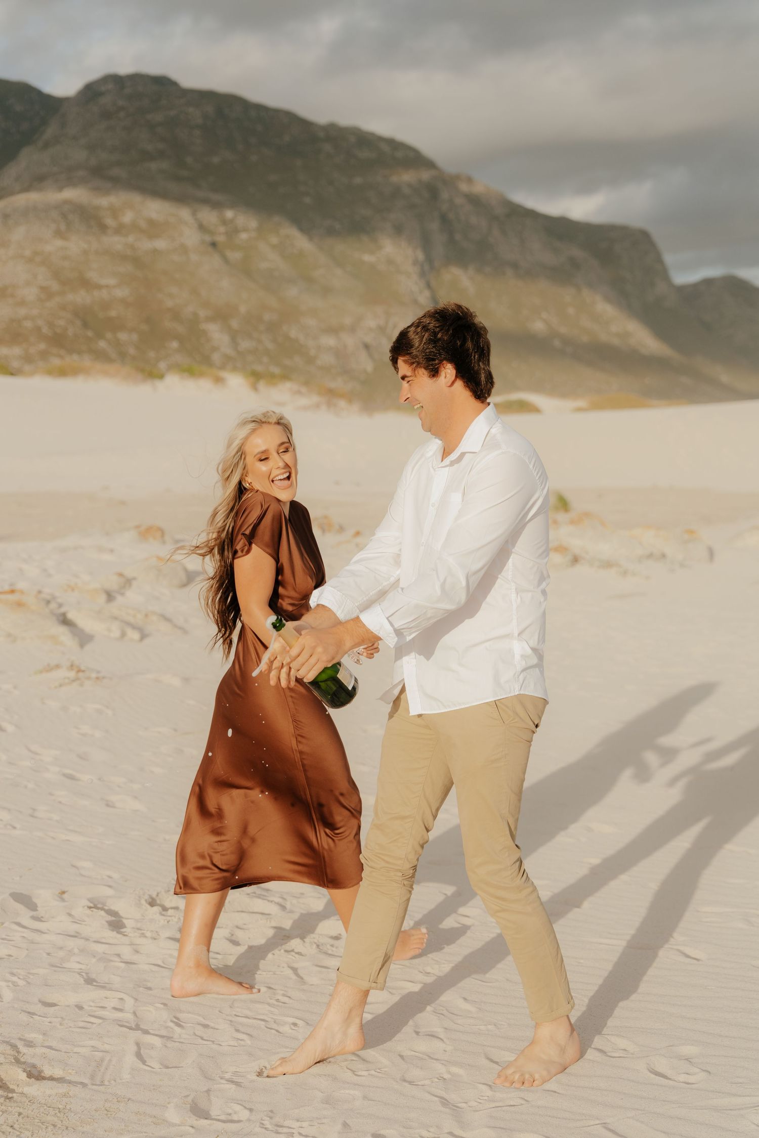 Joyful couple on sandy beach with champagne bottle, woman in brown dress, man in white shirt, mountains behind.