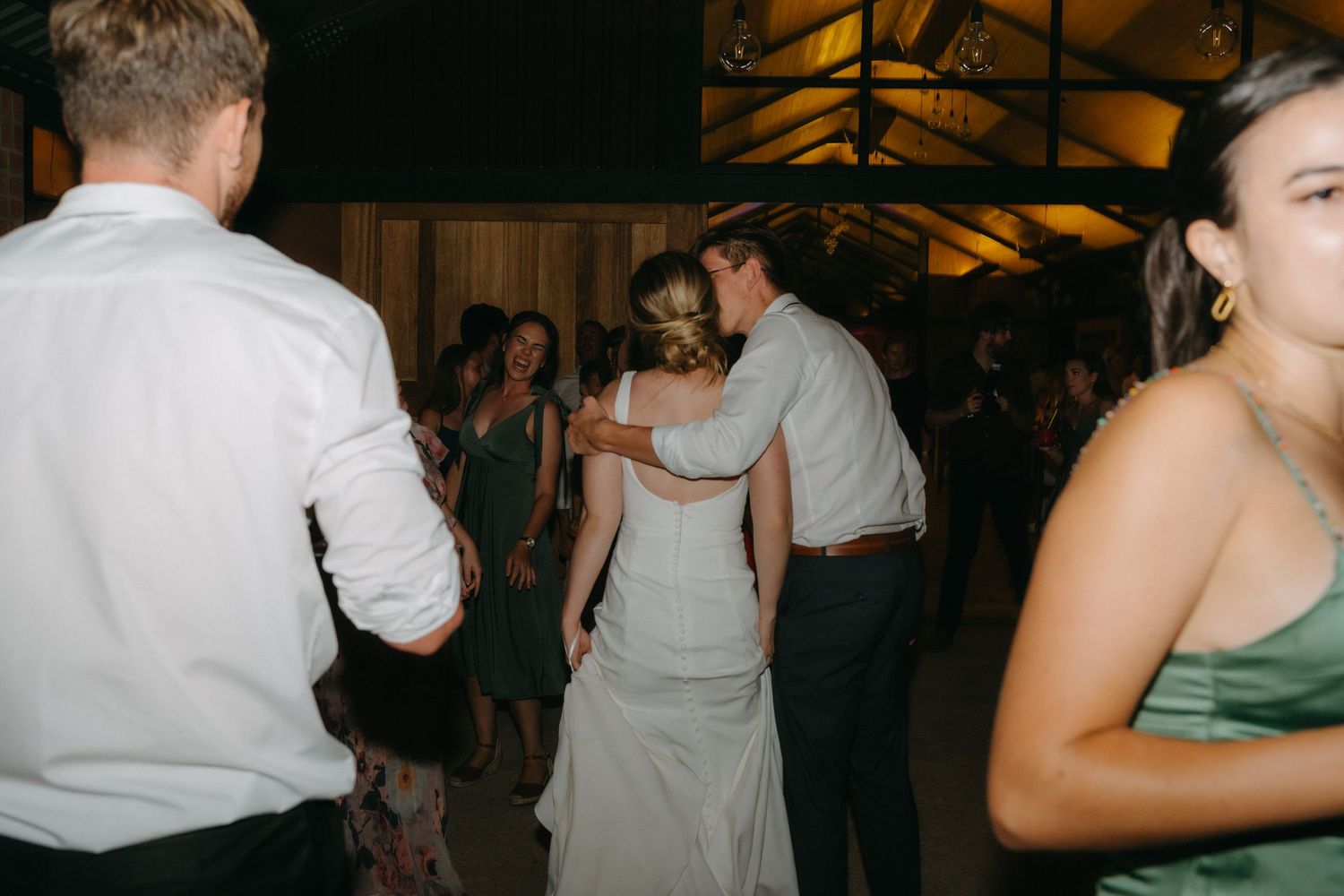 Wedding guests dancing and enjoying themselves on a dimly lit wooden floor at an indoor reception venue.