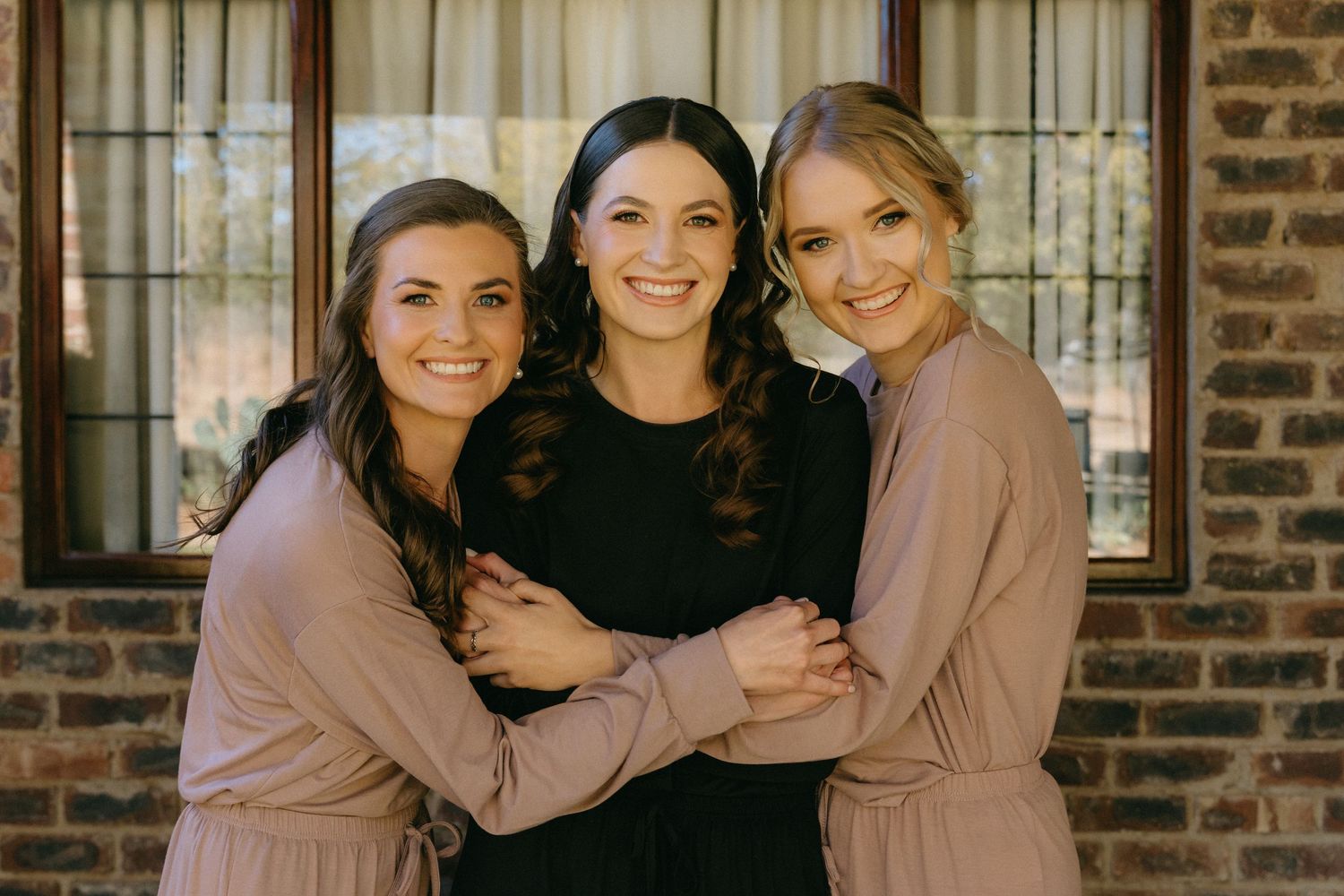 Wedding party members embrace and smile together against a rustic brick wall backdrop.
