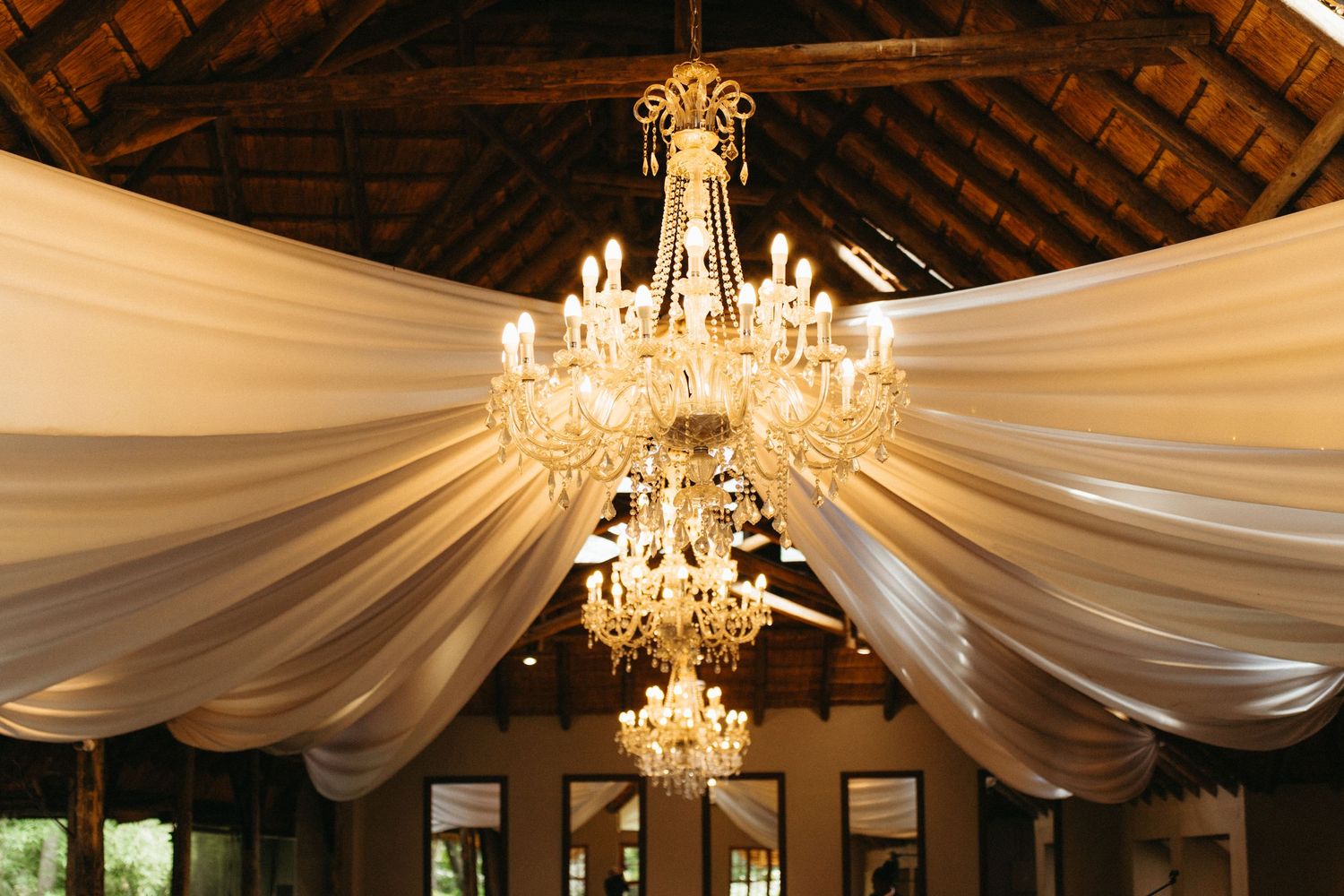 Elegant crystal chandeliers hang from a wooden ceiling draped with flowing white fabric in a reception venue.
