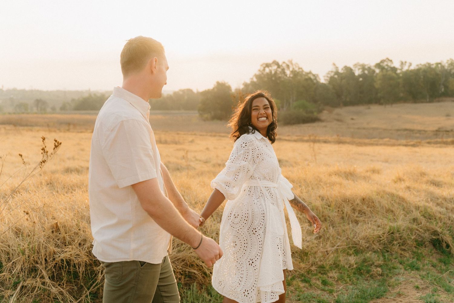 A couple walks hand in hand through a golden field at sunset during a romantic photoshoot.