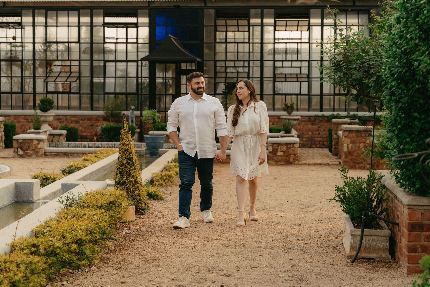 A couple strolls hand in hand through a garden path with rustic brick walls and lush greenery surrounding them.